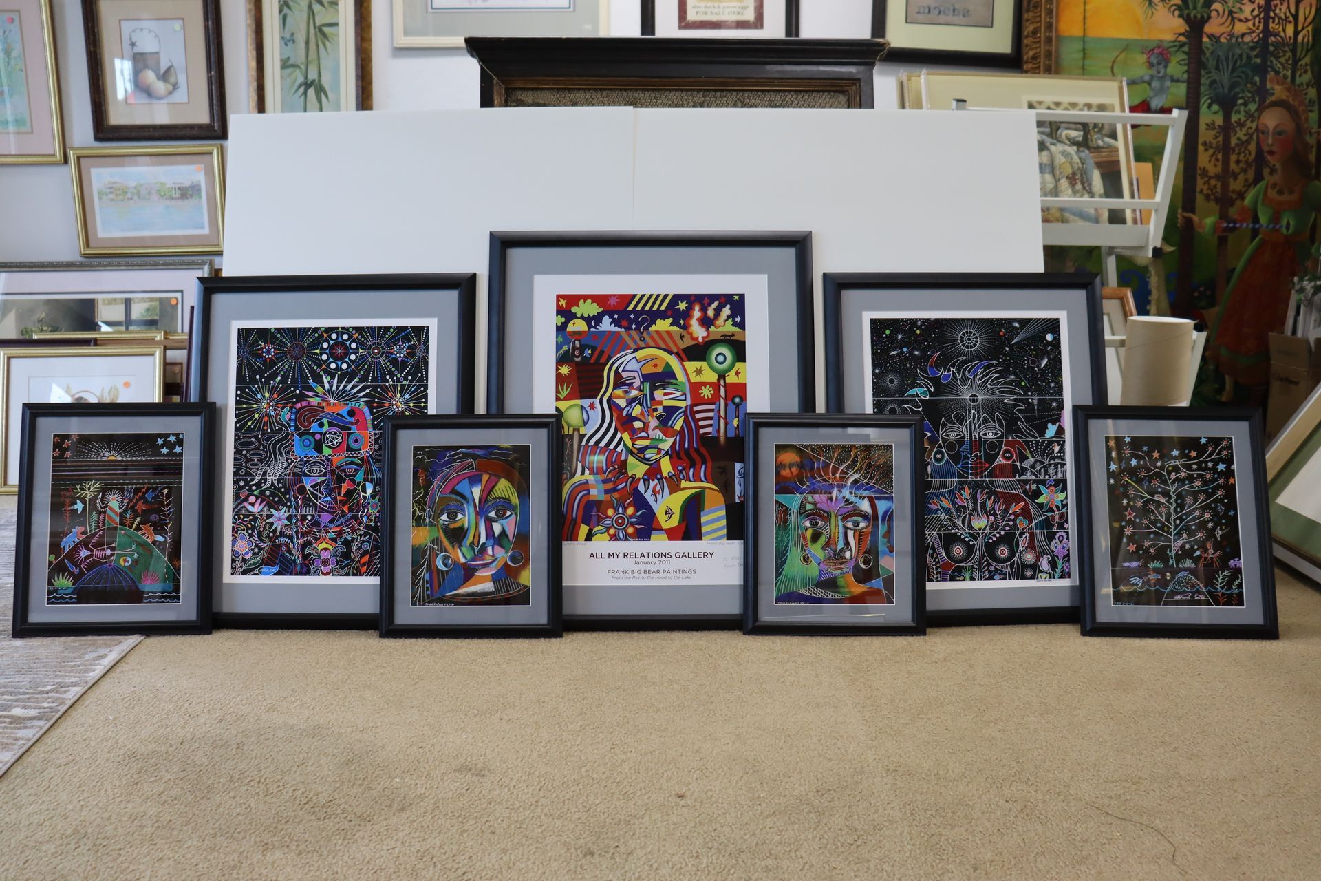 Multiple framed artworks of varying sizes displayed in a row on a floor in front of a white partition.