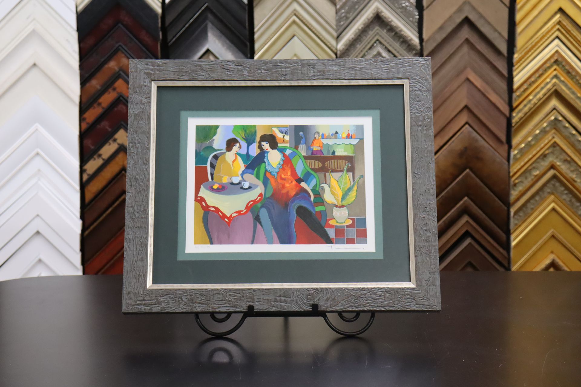 A framed, vibrant painting of two people at a table, centered on a table in front of a display of various picture frames.