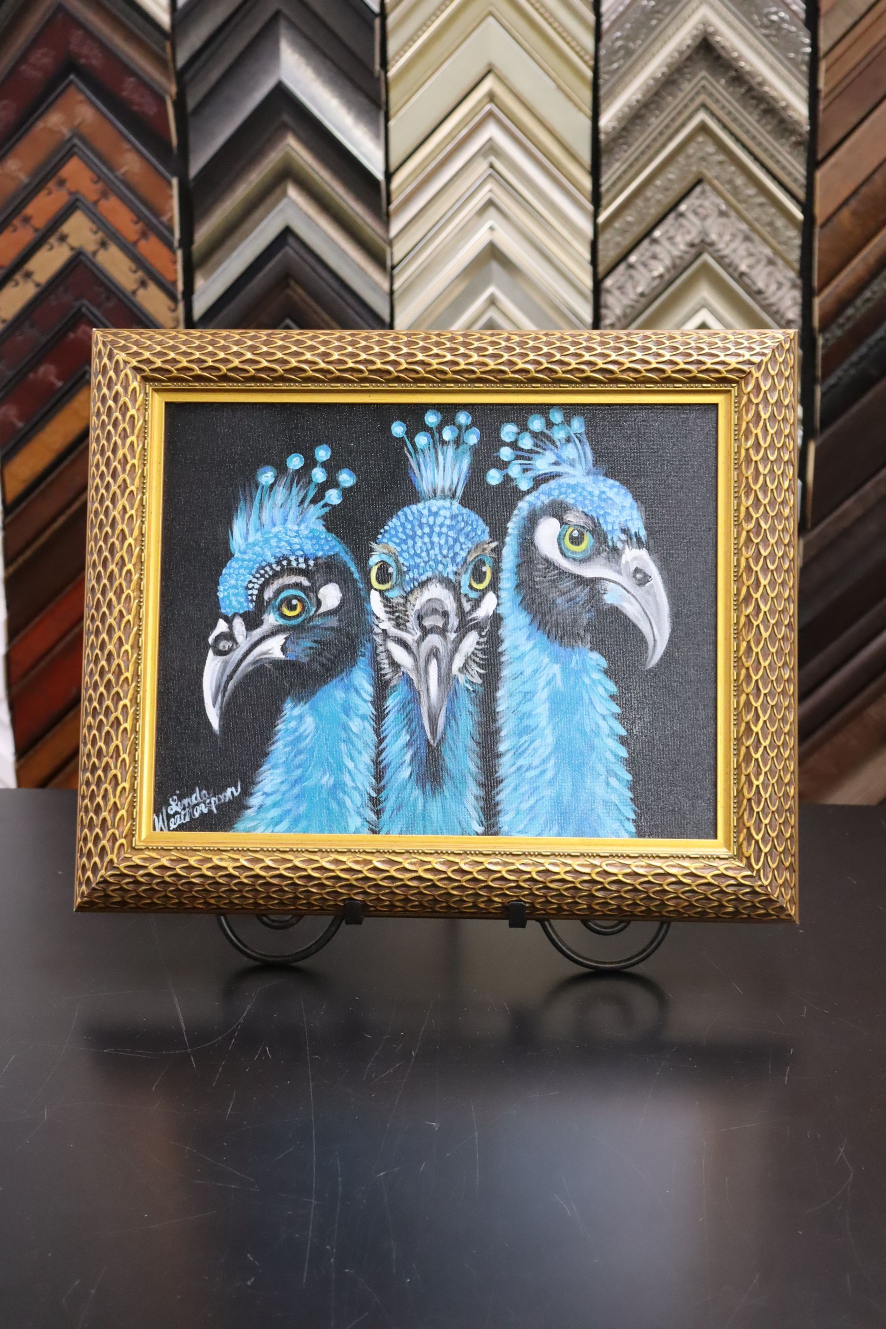 A textured, gold-framed painting of three blue peacock heads on a dark, speckled background, sitting on a display stand.