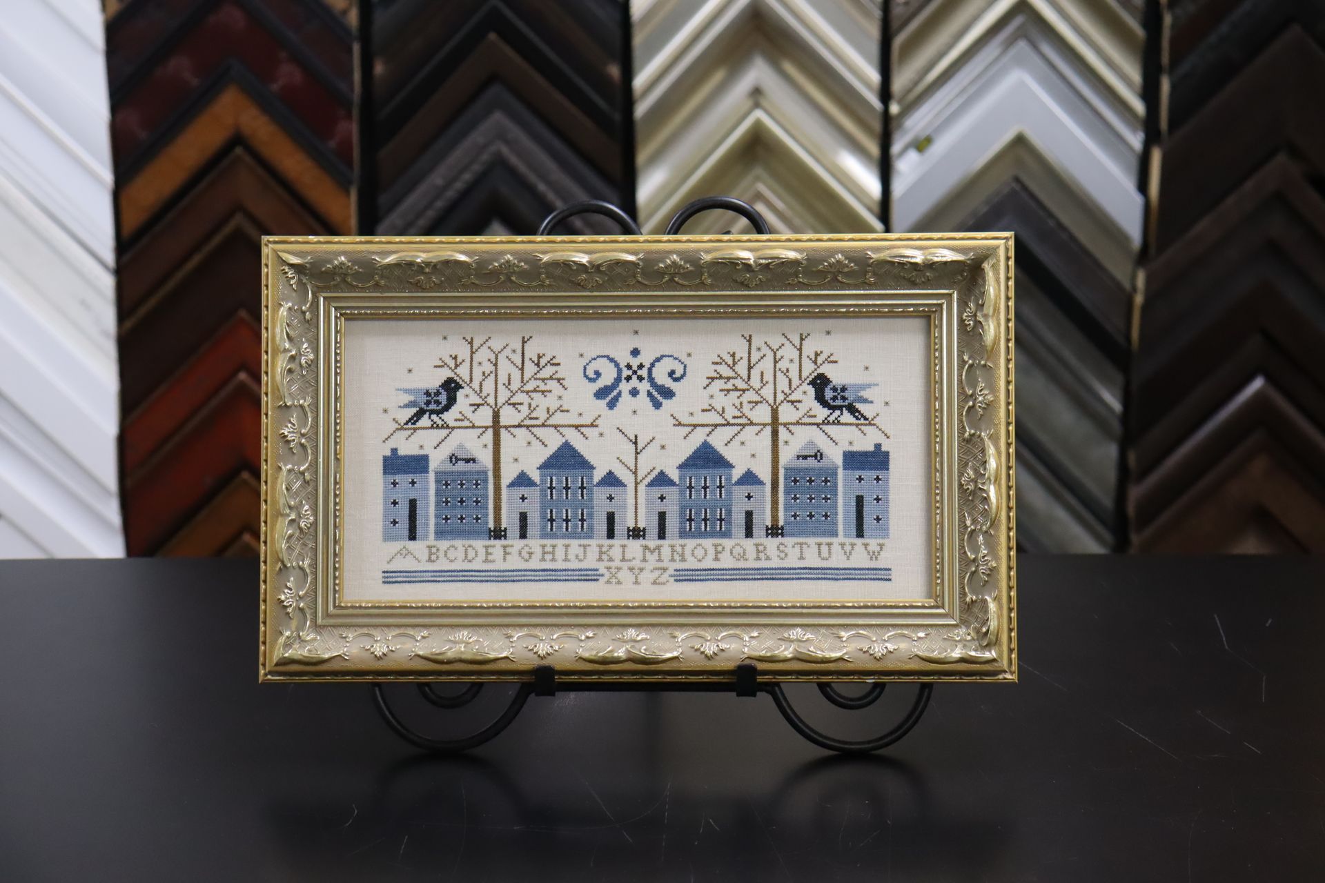 A framed needlepoint sampler with blue bird, tree, and house designs sits on a metal stand against a background of frames.