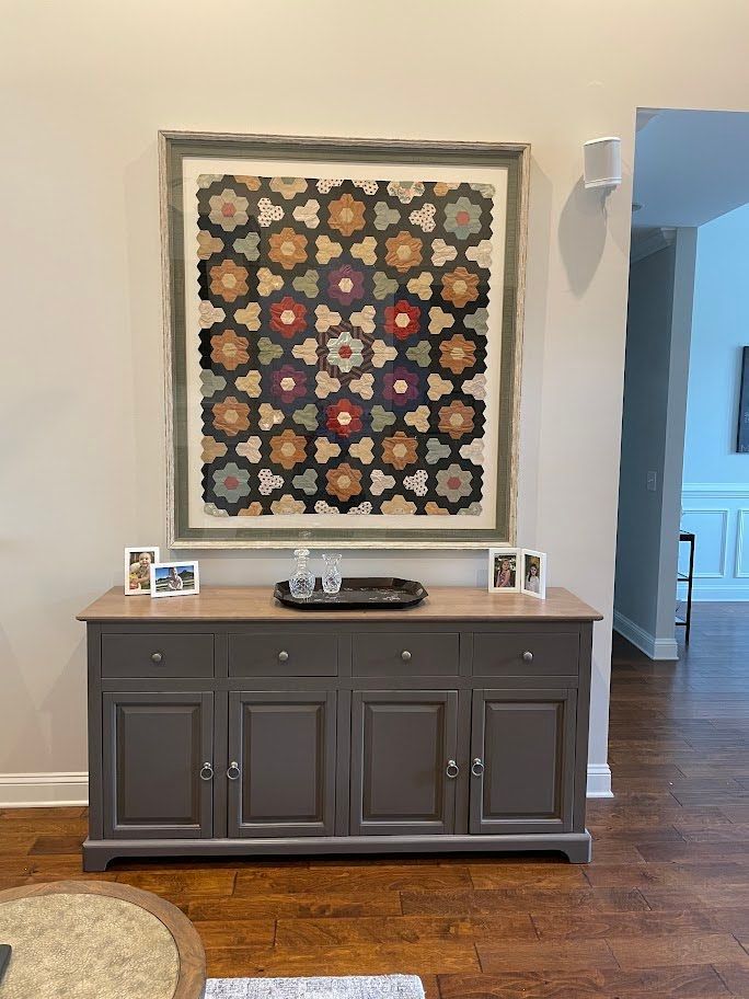 A framed geometric patterned quilt hangs above a dark gray, four-door sideboard with a wooden top in a home setting.