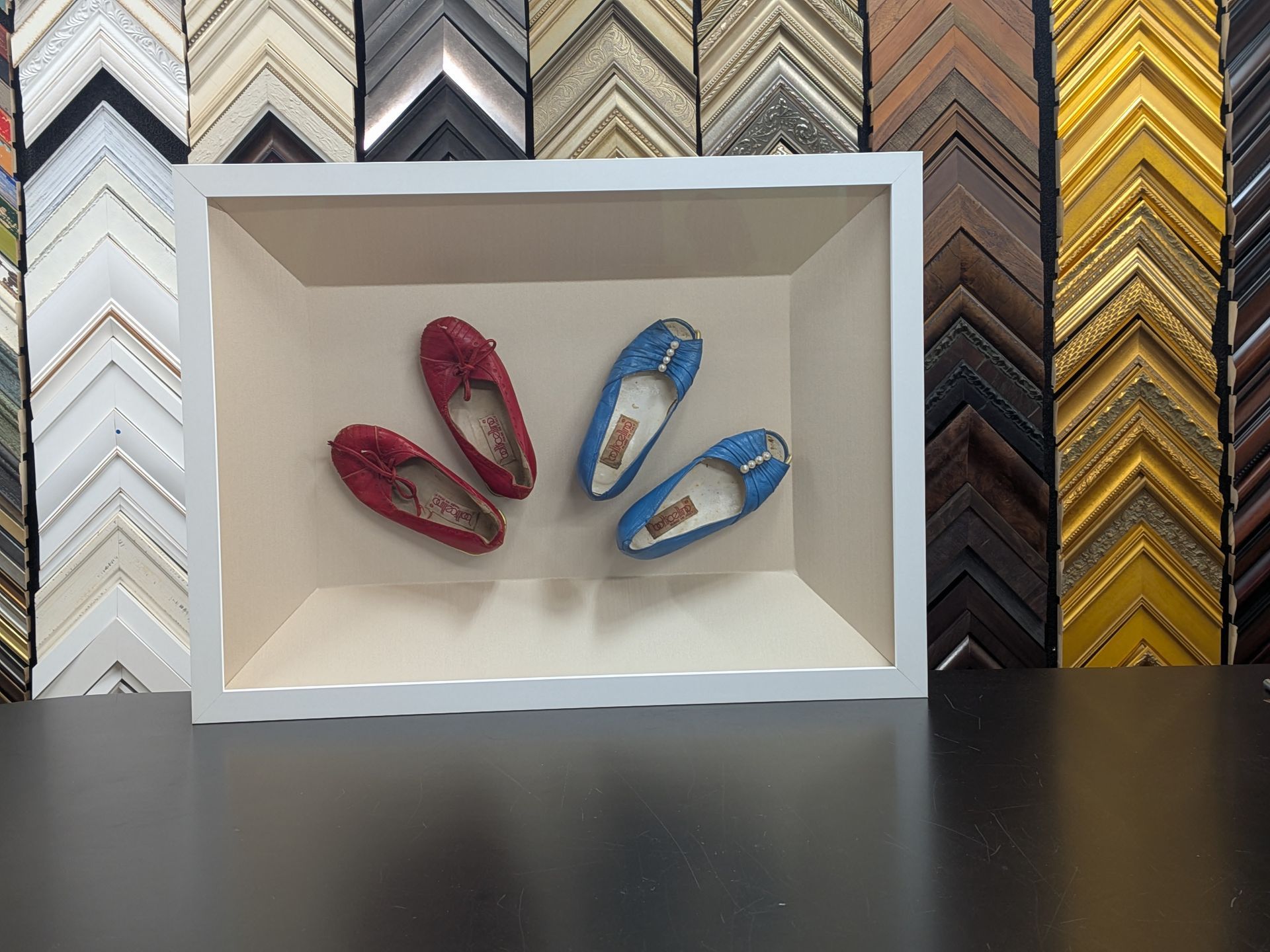 Two pairs of small, colorful shoes in a white shadow box, displayed against a wall lined with various picture frames.