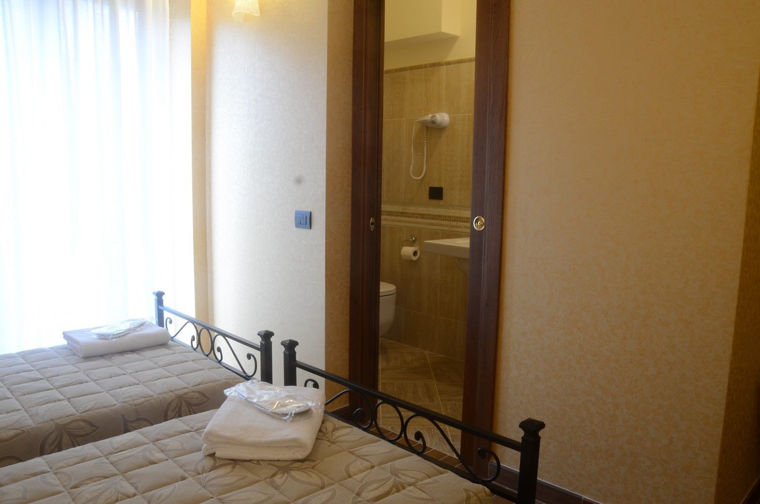 Piccola camera da letto con due letti singoli, tende bianche e porta del bagno aperta.