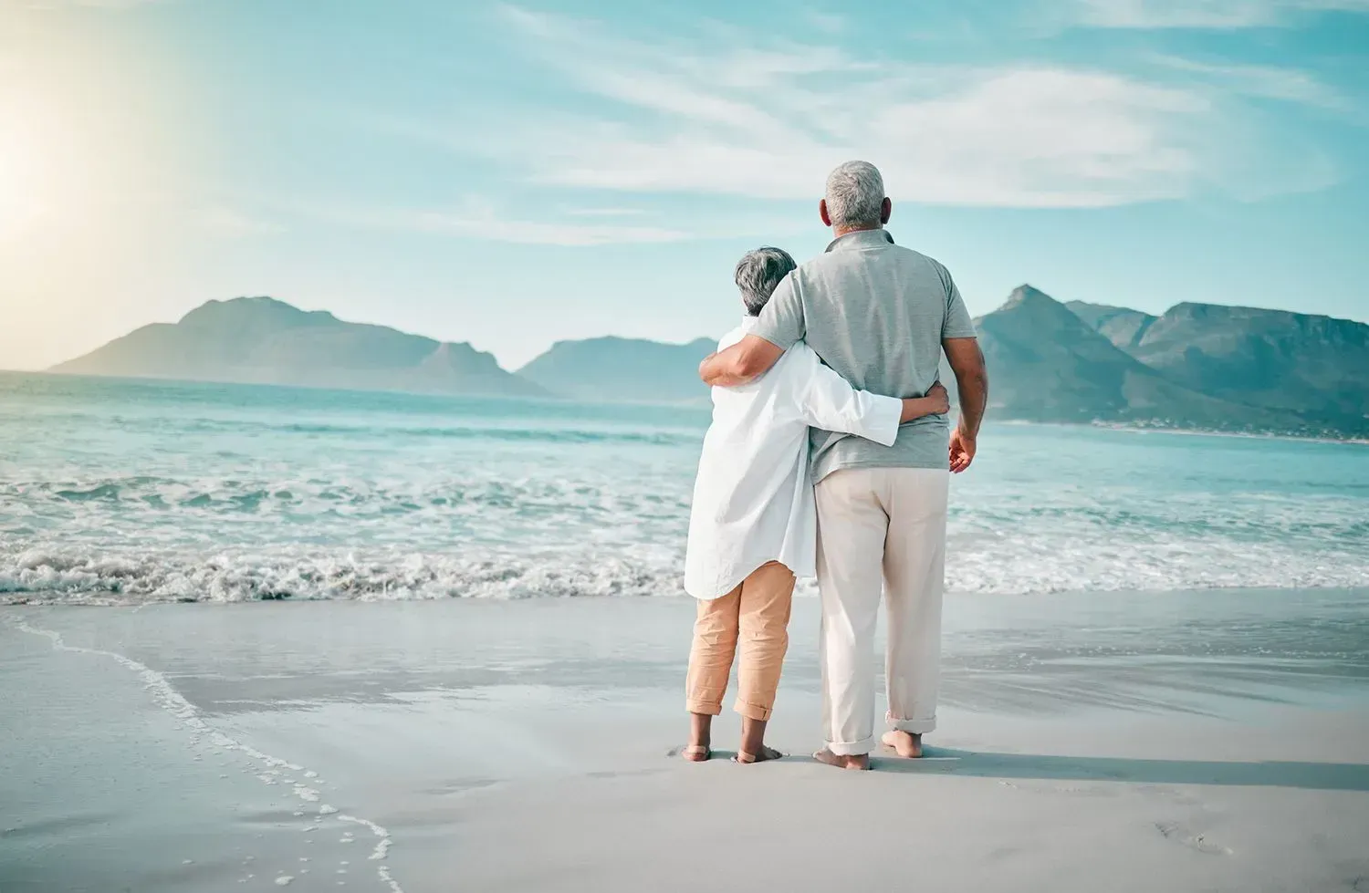 An elderly couple is standing on a beach looking at the ocean.