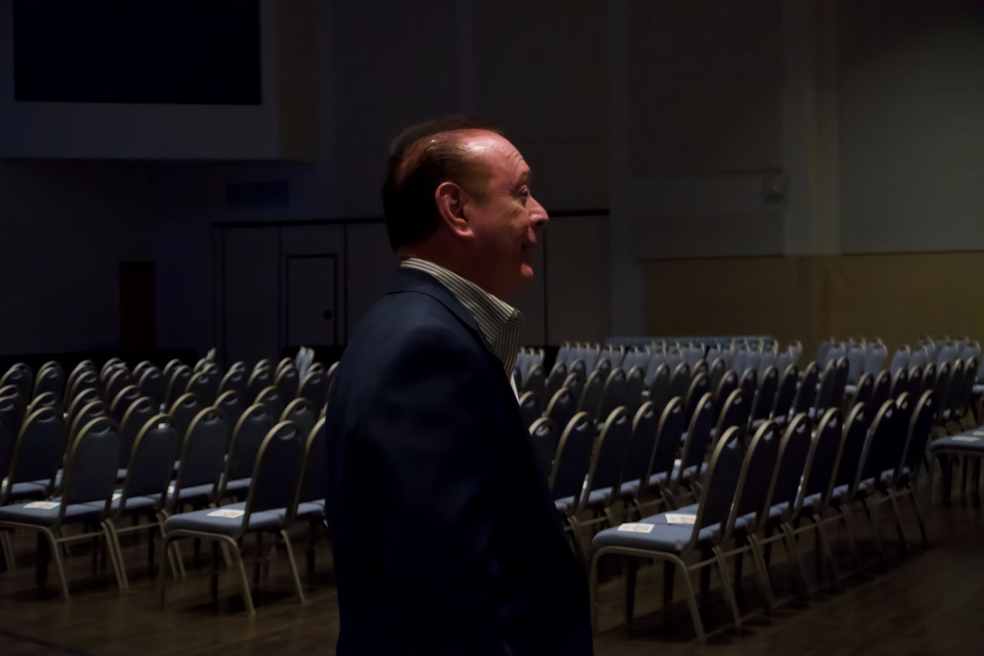 A man in a suit is standing in front of rows of chairs in a dark room.
