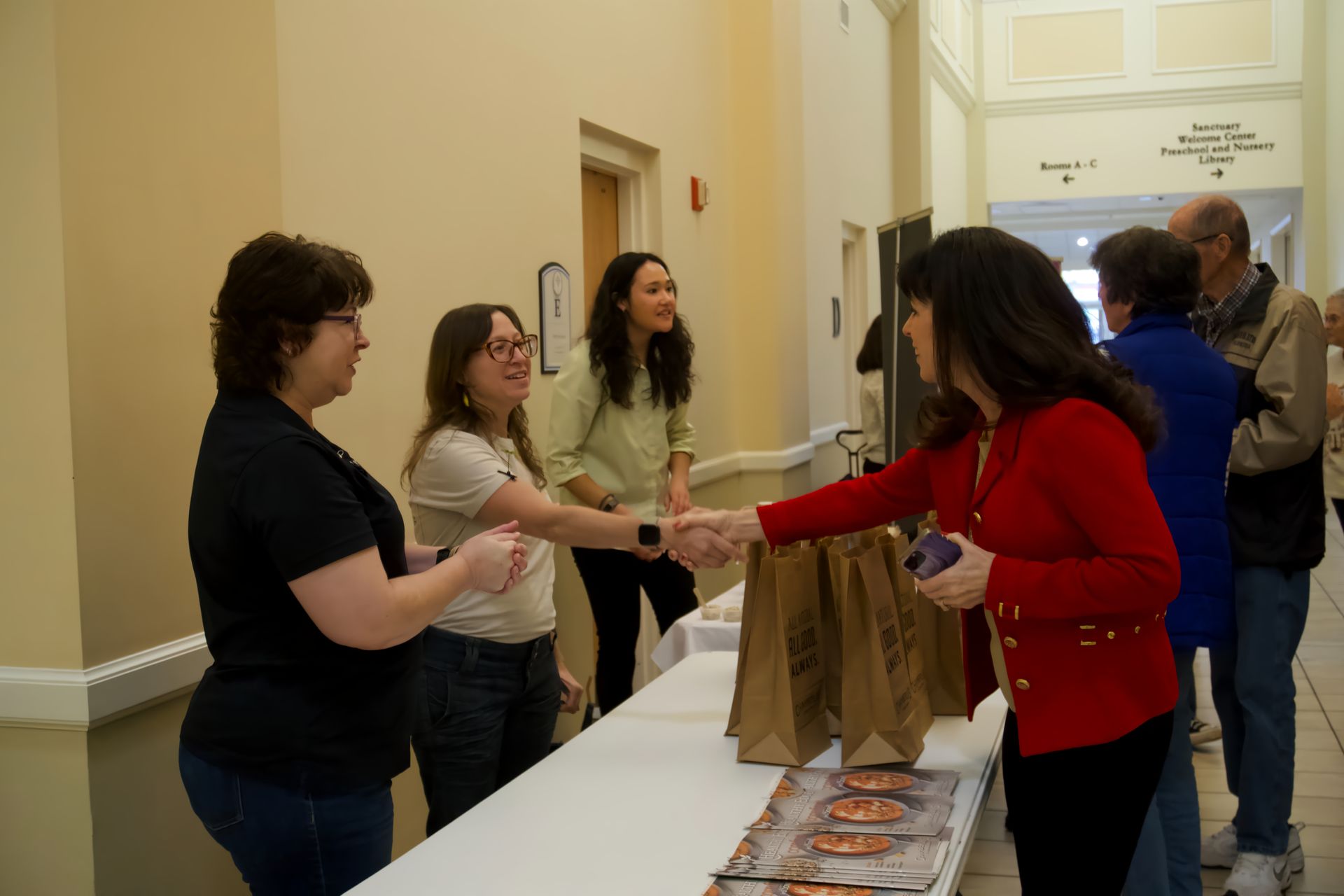 A woman in a red jacket is shaking hands with another woman at a table.