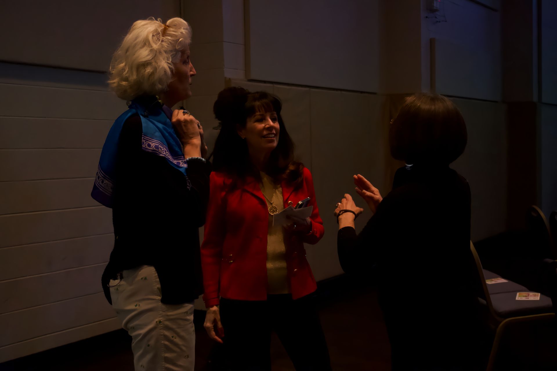 Three women are standing next to each other in a dark room talking to each other.