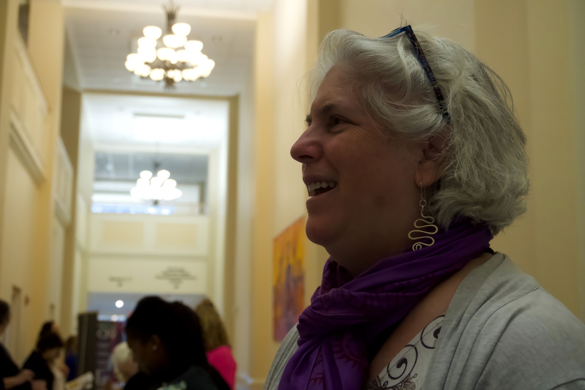 A woman wearing a purple scarf and earrings smiles in a hallway