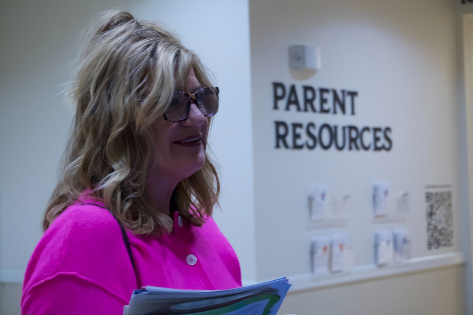 A woman in a pink shirt is standing in front of a sign that says parent resources.
