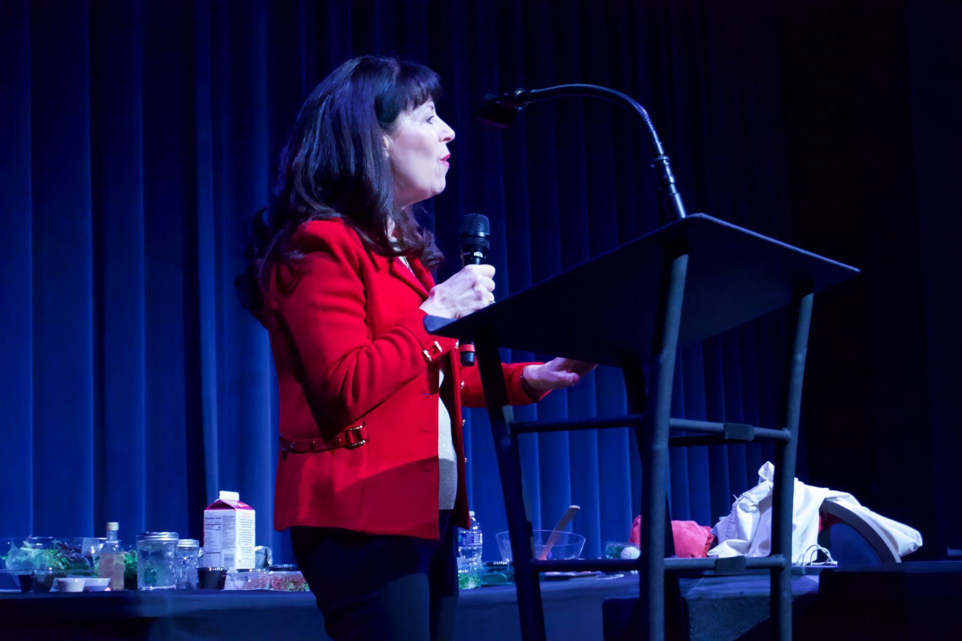 A woman in a red jacket is standing at a podium holding a microphone.