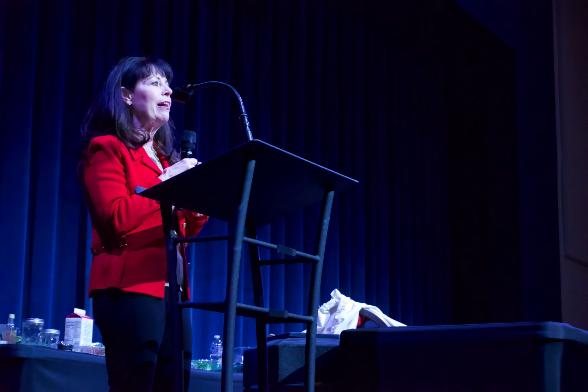 A woman in a red jacket is standing at a podium speaking into a microphone.