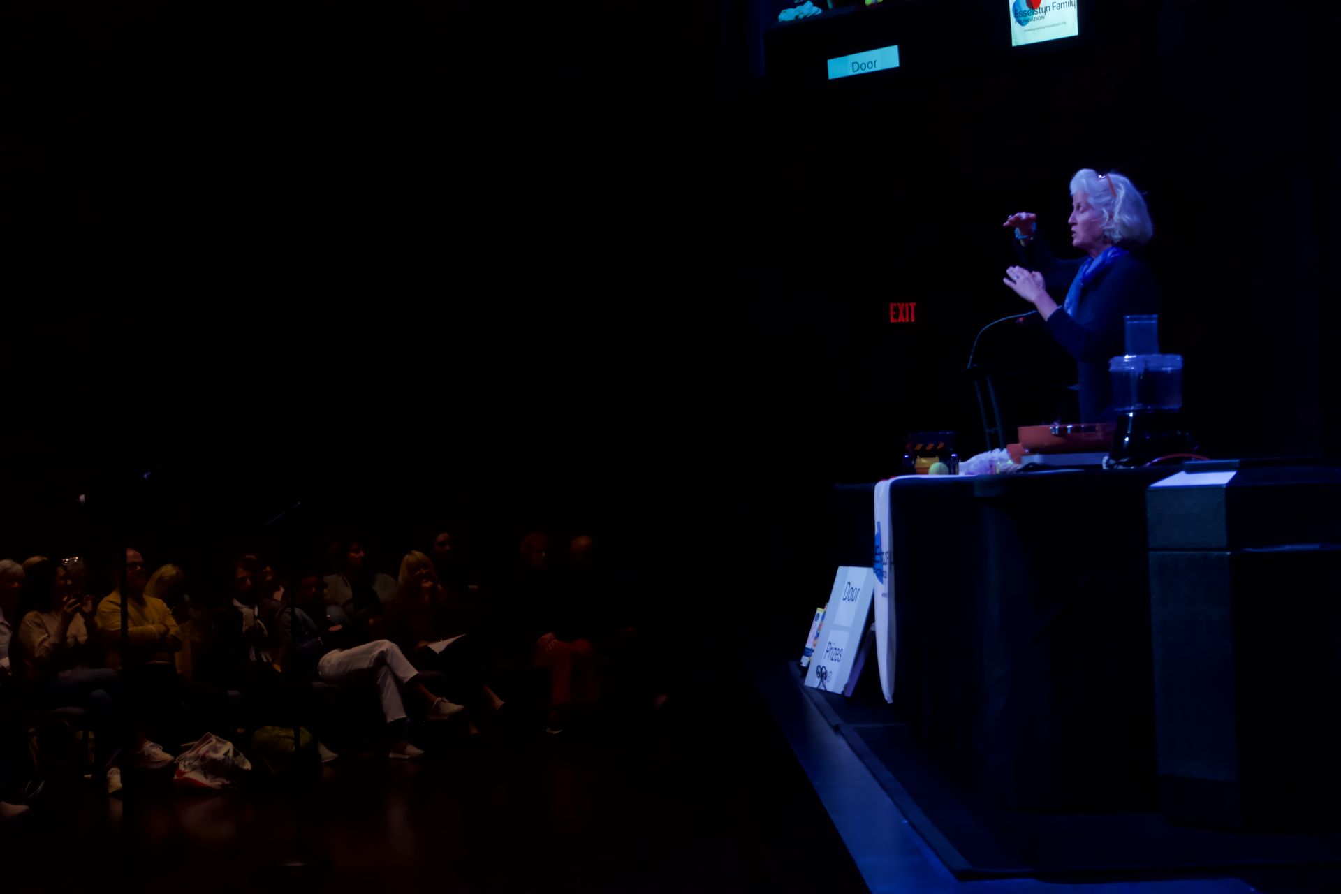 A woman is giving a speech in front of a crowd in a dark room.