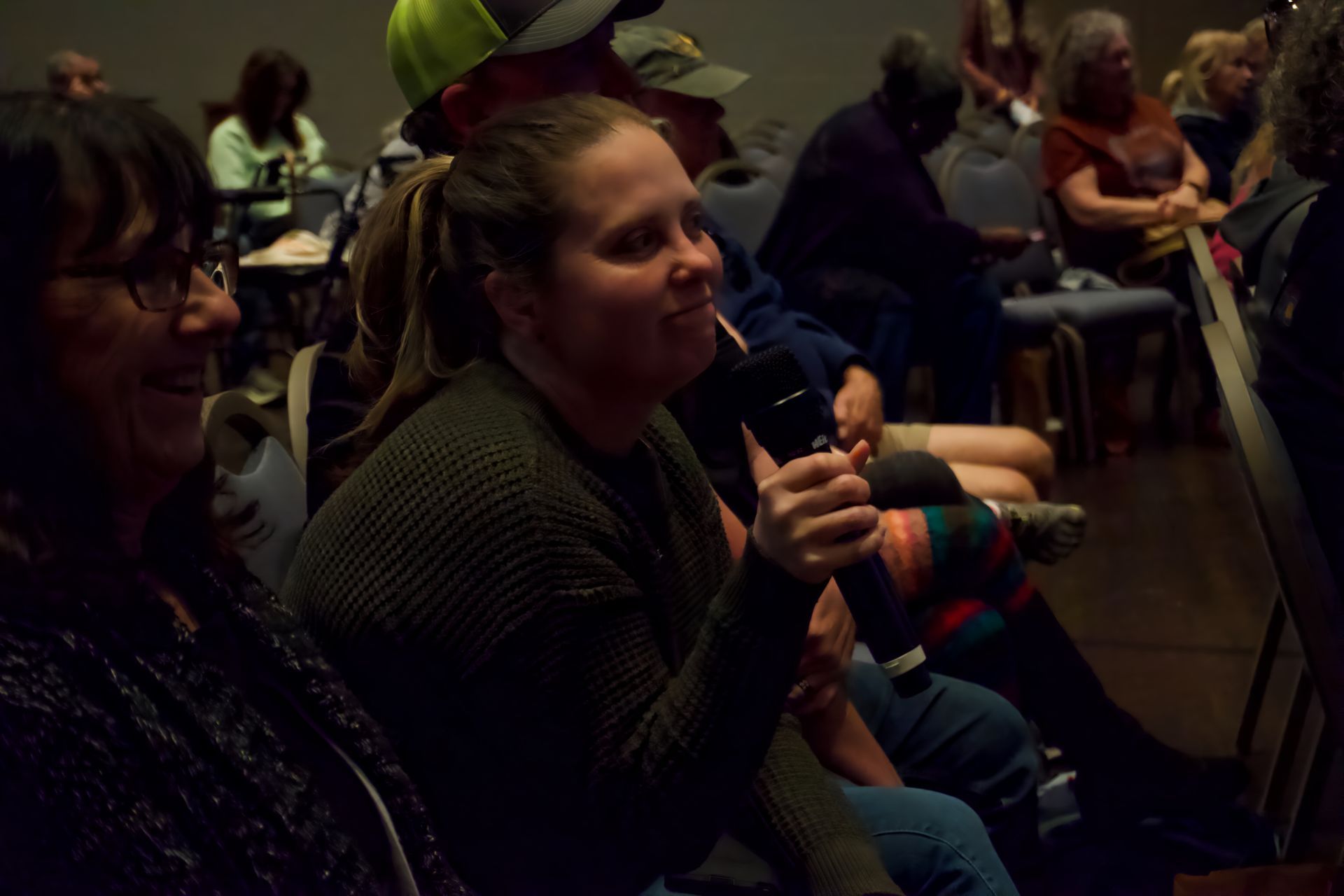 A woman is sitting in a crowd of people holding a microphone.