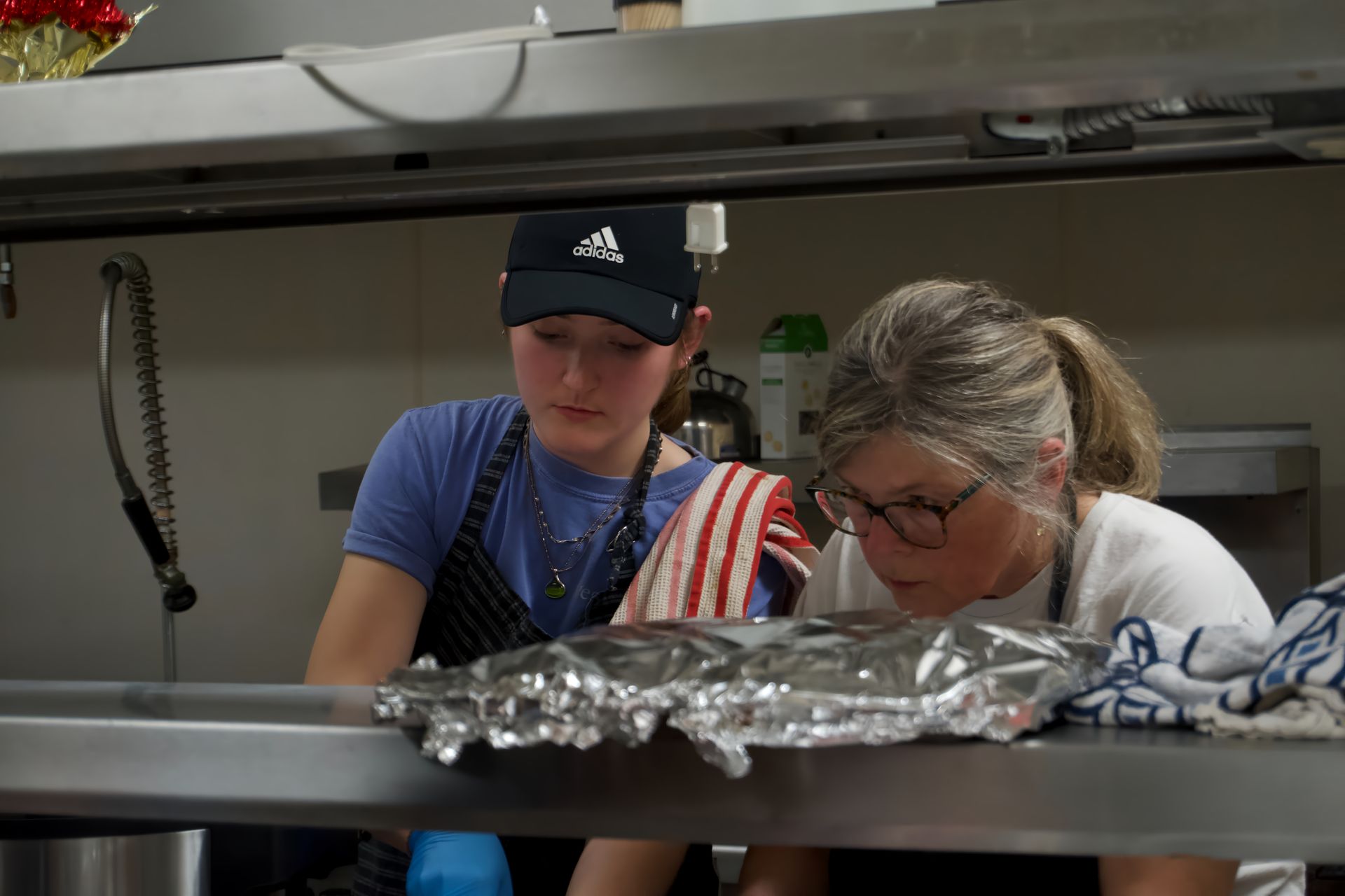 Two women are working in a kitchen and one of them is wearing an adidas hat.