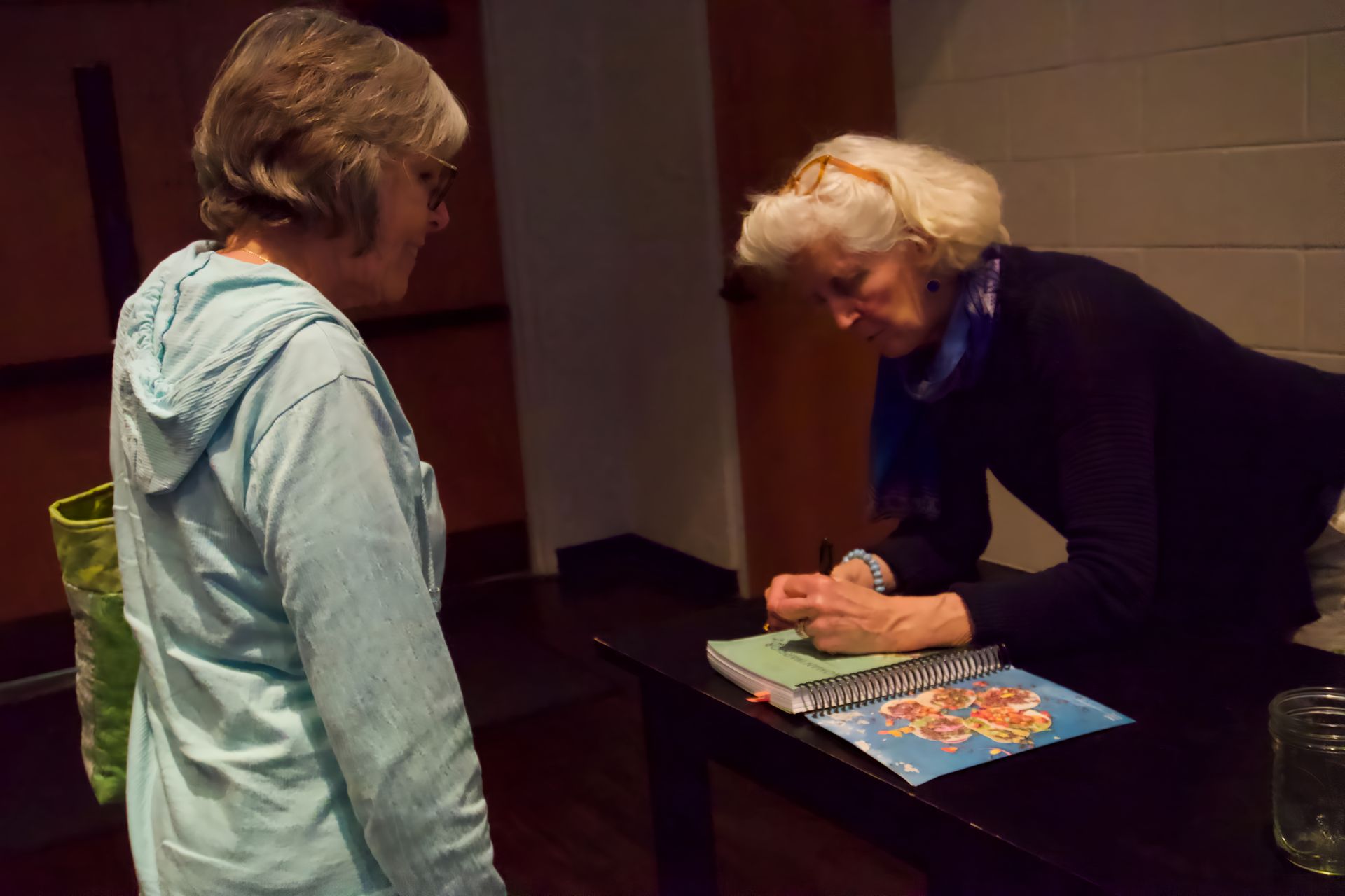 A woman is signing a book for another woman at a table.