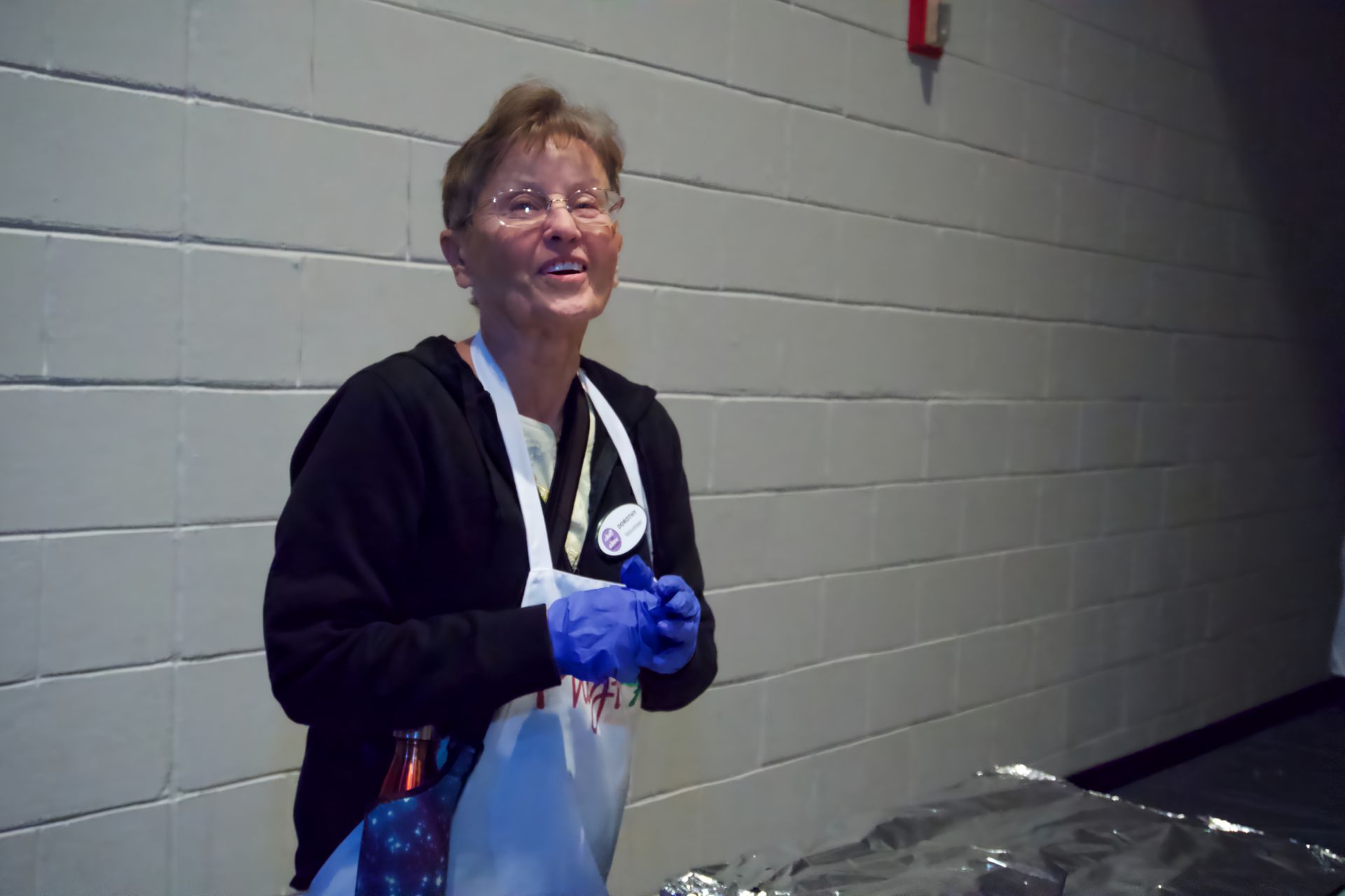 A woman wearing an apron and blue gloves stands in front of a brick wall