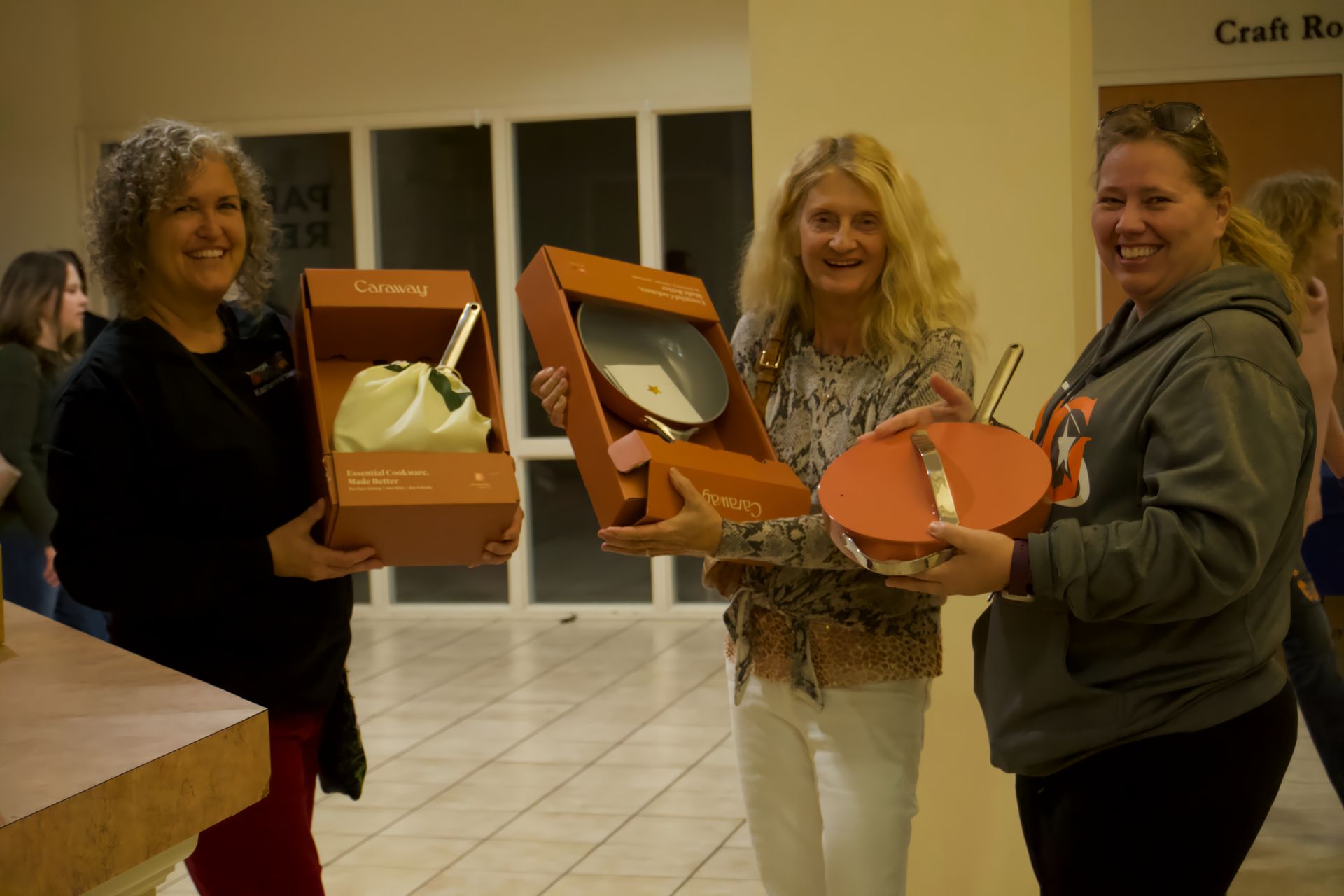 Three women are holding boxes in front of a sign that says craft room