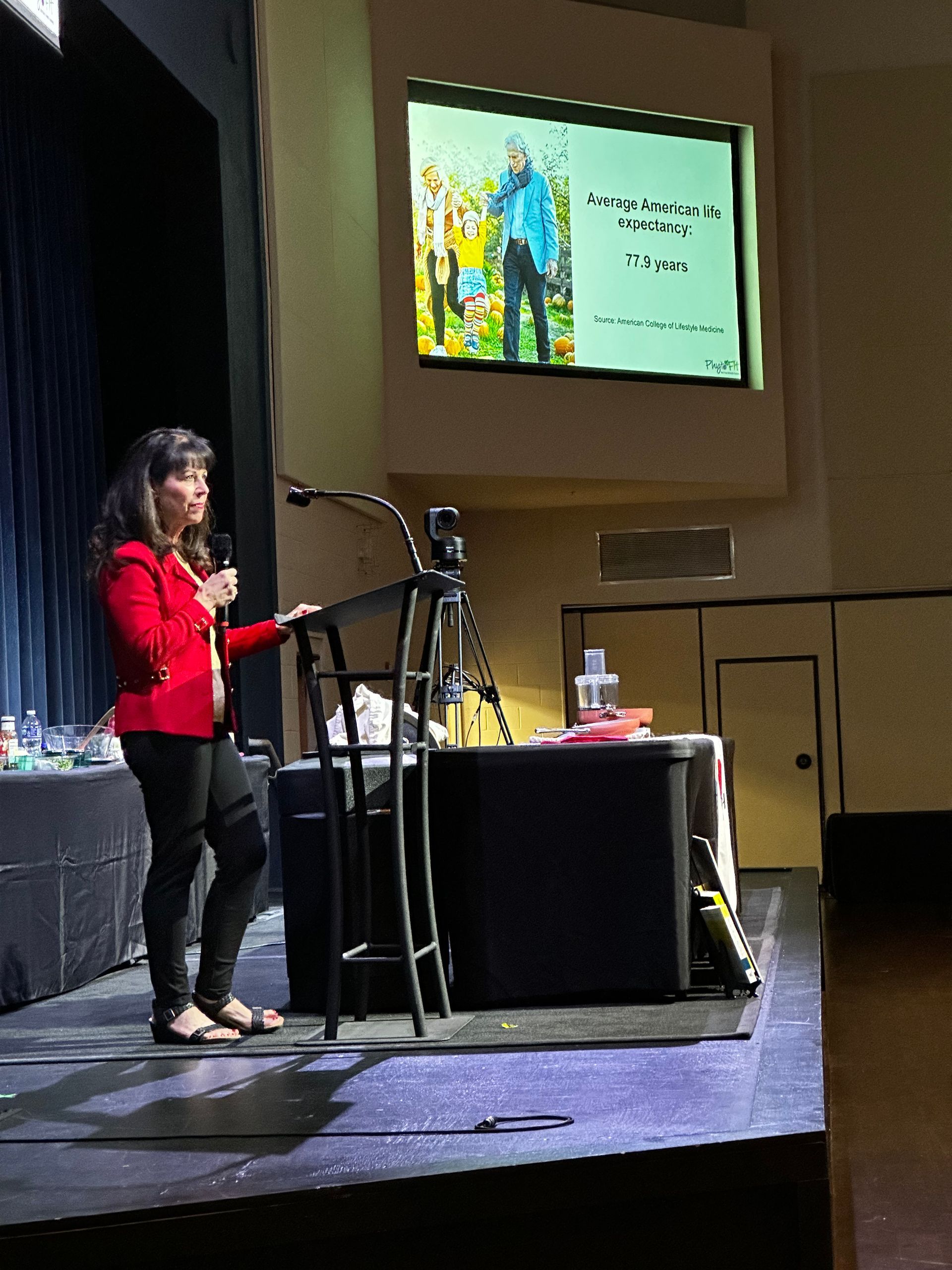 A woman in a red jacket is giving a presentation on stage