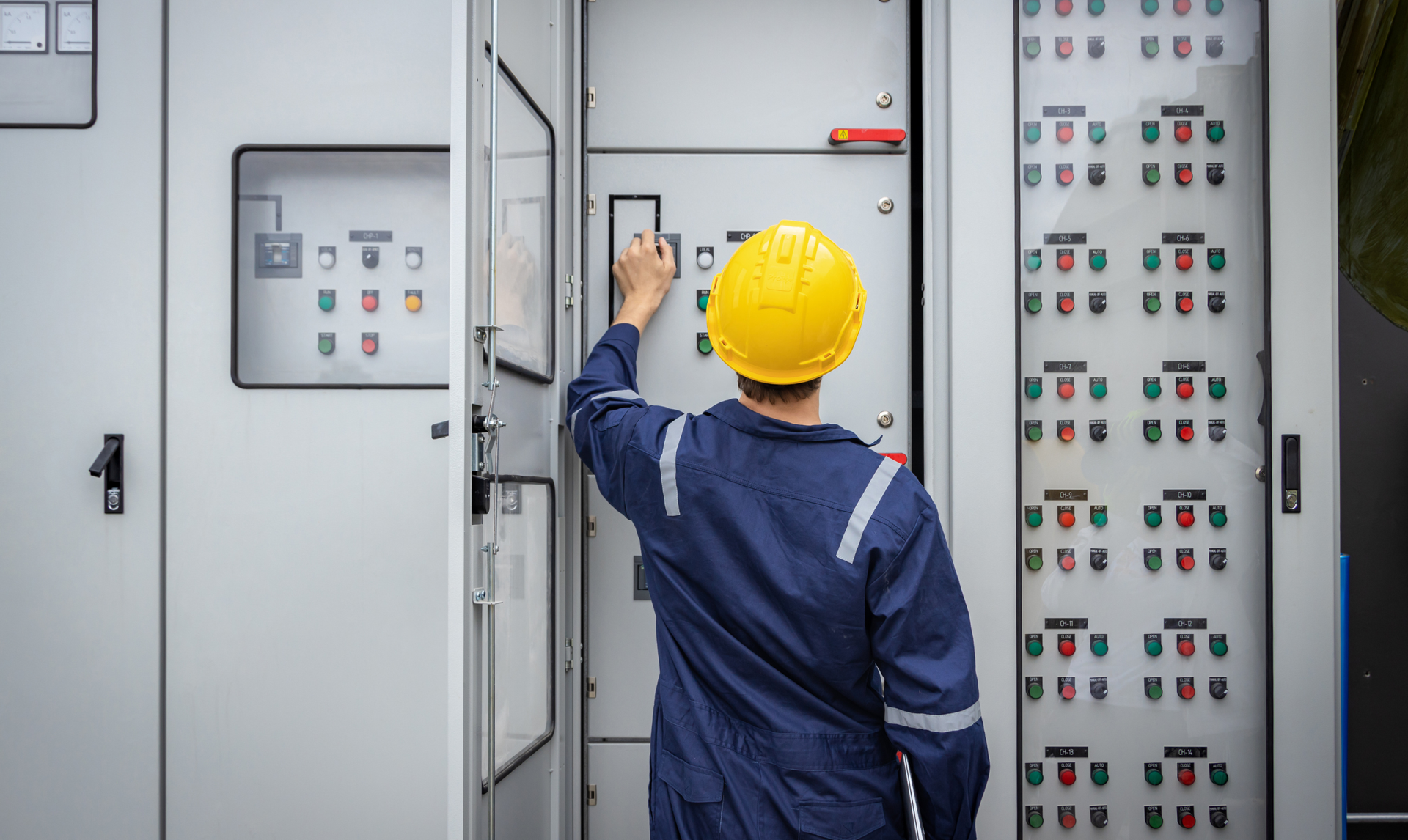 Electrician in blue uniform and yellow hard hat working on electrical panel.