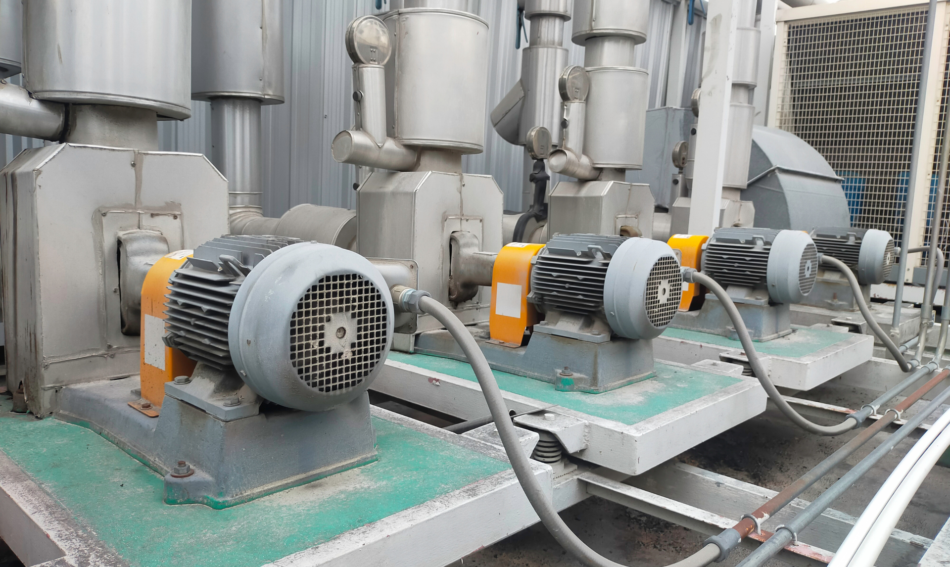 Three industrial pumps with electric motors, metal and silver tones, on green platforms, with gray cabling.
