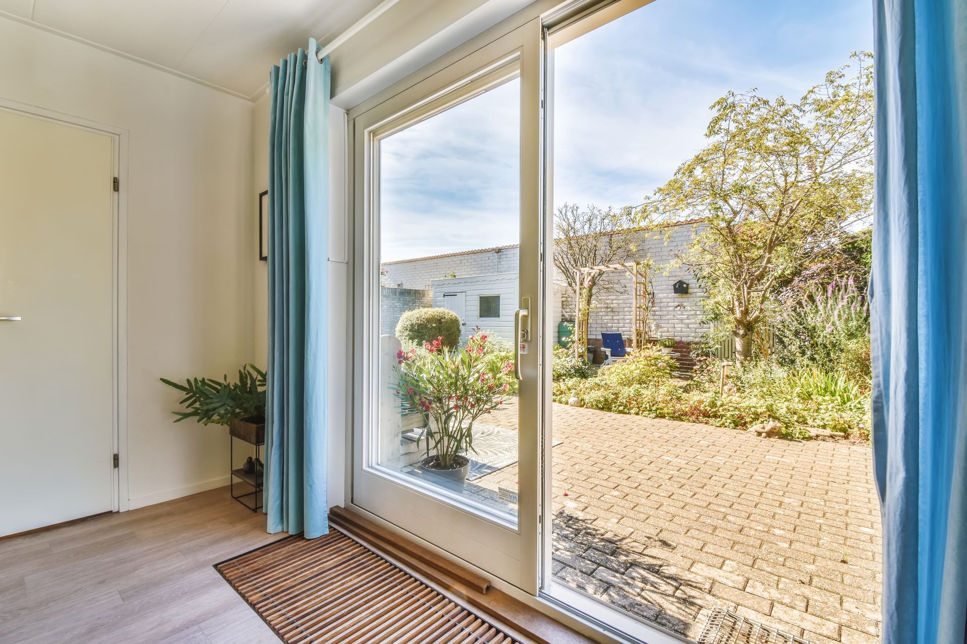 Bright room with open sliding glass door, blue curtains, and a sunlit garden patio outside