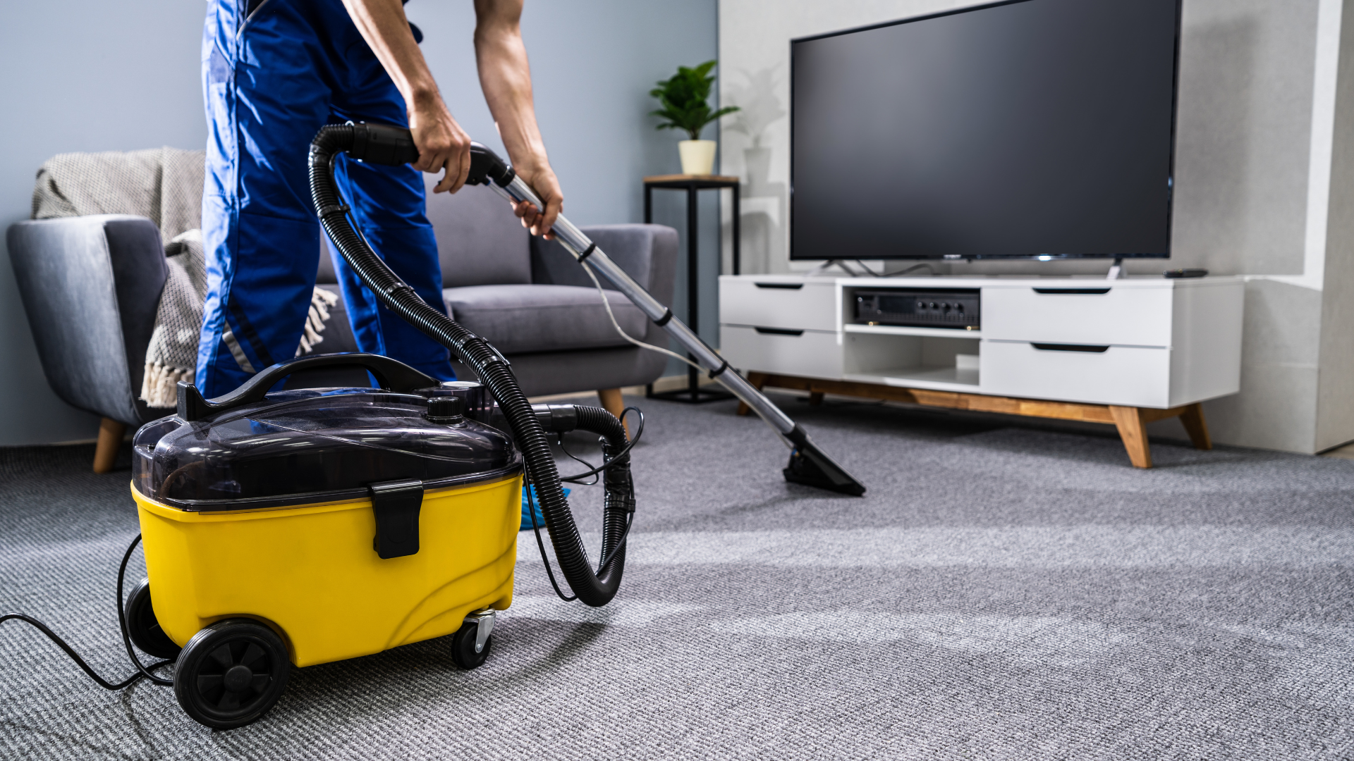 A person in blue work clothes uses a yellow canister vacuum cleaner to clean a grey carpet in a living room.
