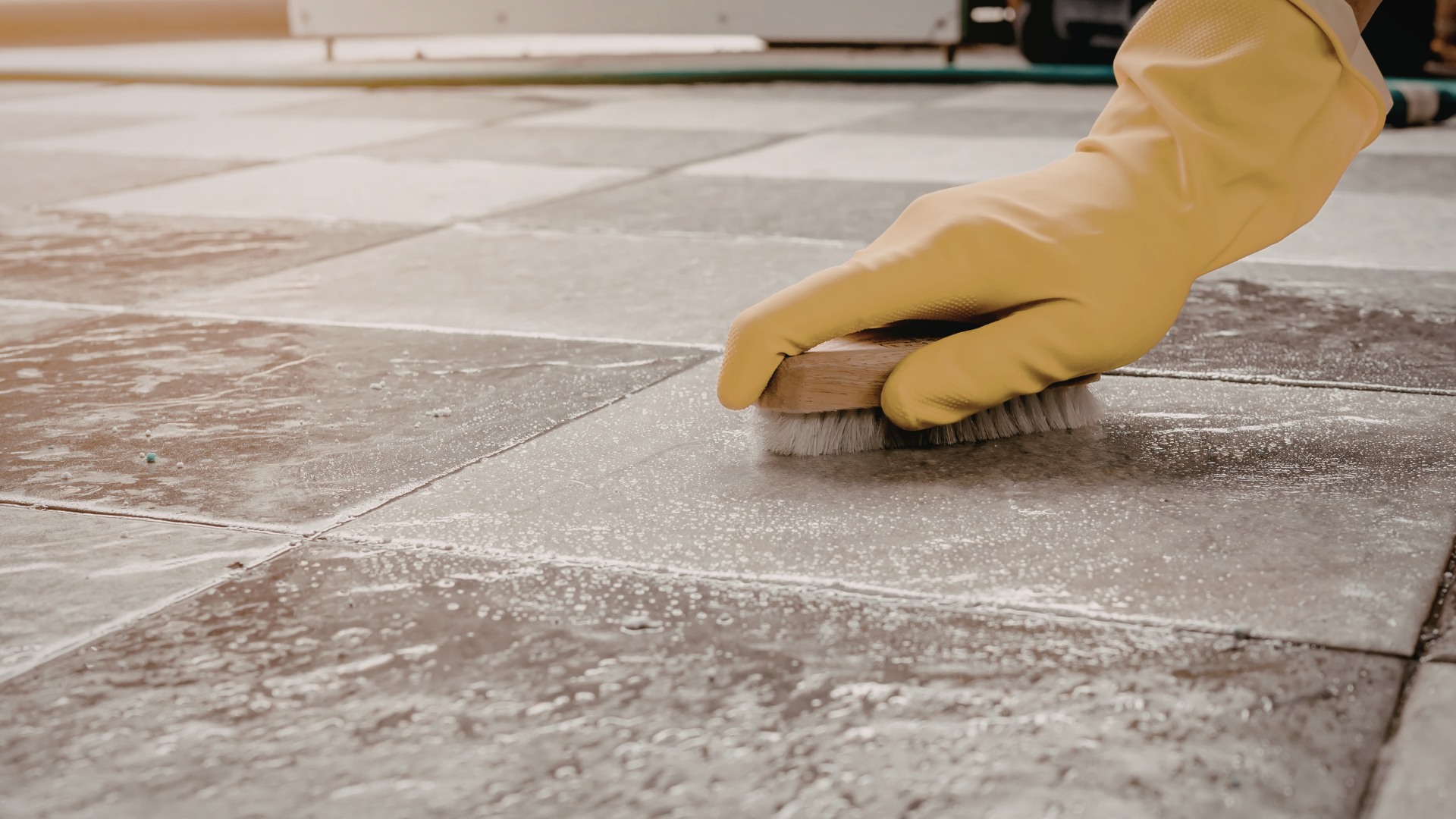 A gloved hand uses a scrub brush to clean light-colored, patterned floor tiles.