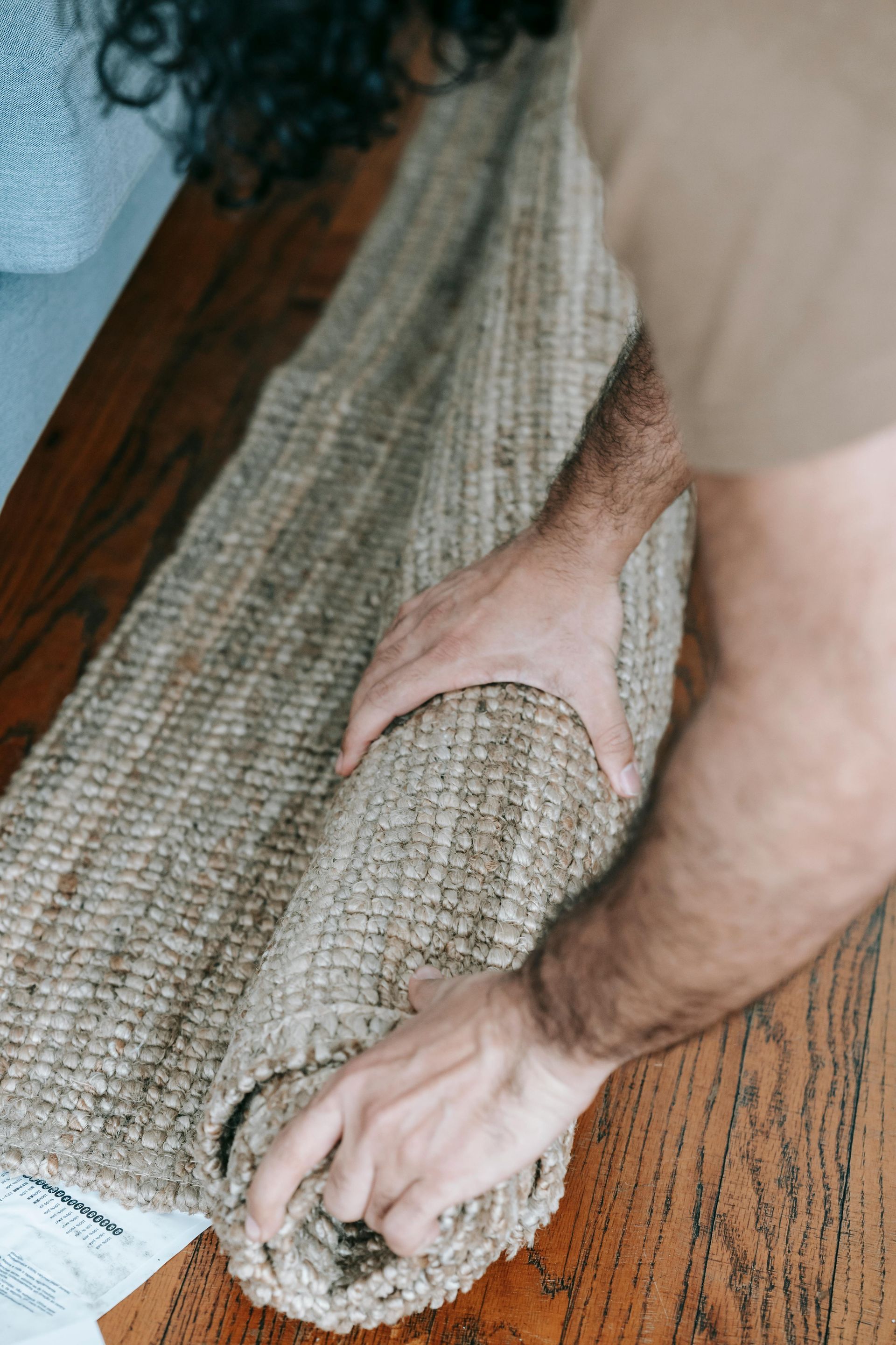 A person rolling up a textured, natural fiber rug on a wooden floor.
