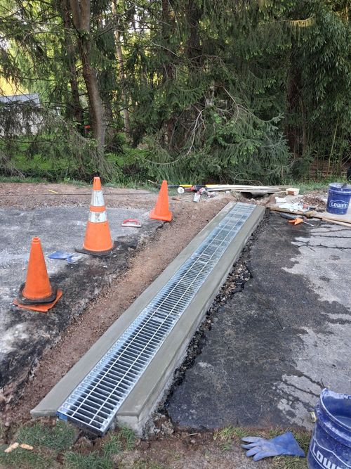 Roadside drainage trench with metal grating, orange cones, and construction debris along a paved path