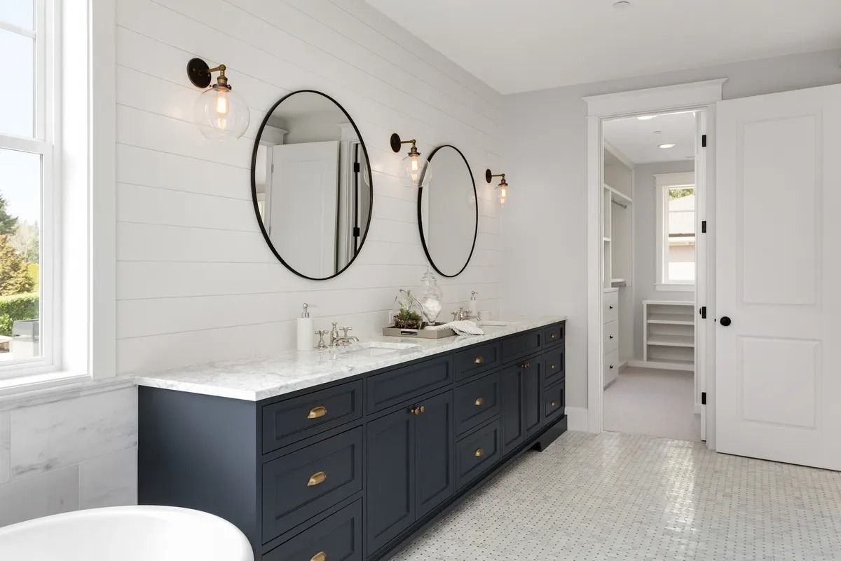 Bright bathroom with navy vanity, round mirrors, white tile walls, and a doorway to a shower area.