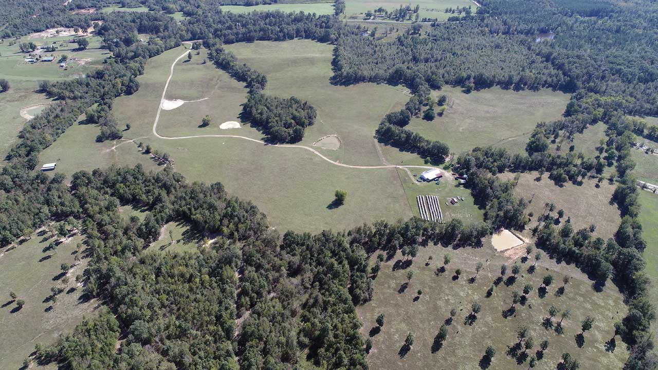 Aerial view of a rural landscape with fields, trees, and scattered buildings under a blue sky.