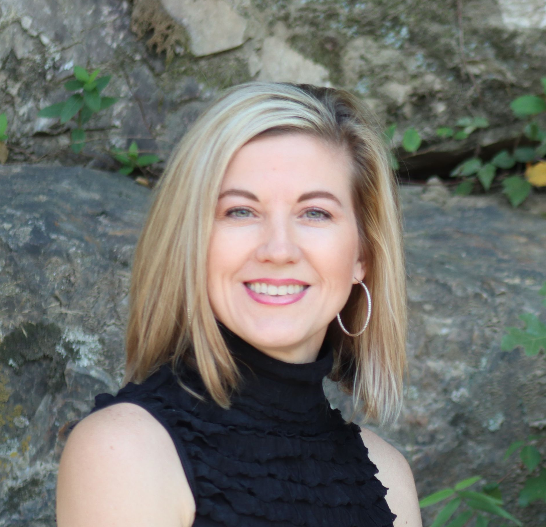 Woman with blonde hair smiles, wearing a black top, in front of a stone wall.