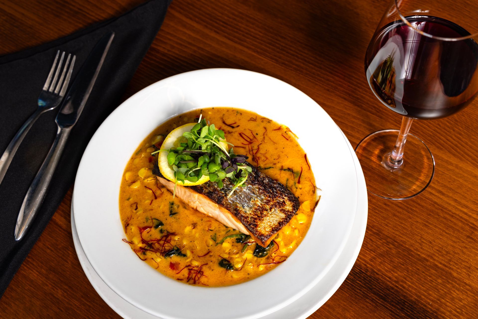 Plate of fish with sauce, garnish, and a glass of red wine on a wooden table with silverware.
