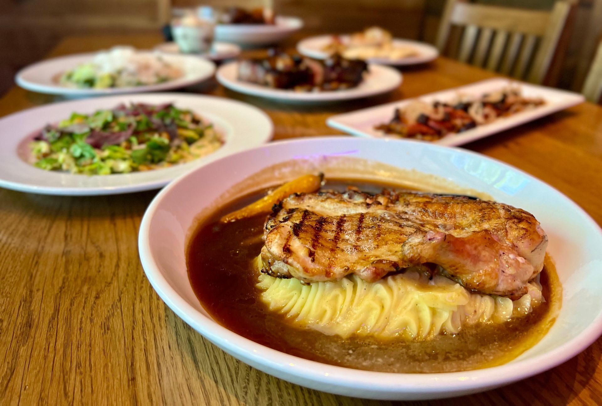 A white plate with pork chop on mashed potatoes, other dishes on a wooden table.