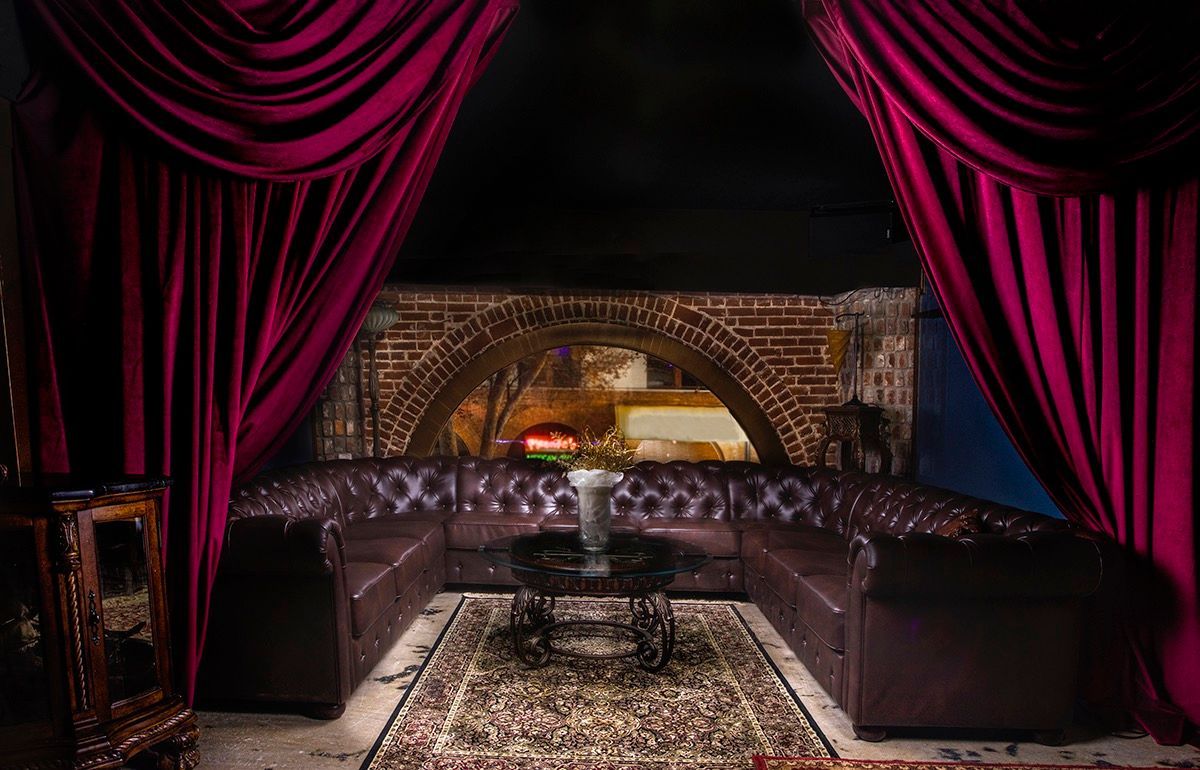 Luxurious lounge with burgundy drapes, leather sofa, ornate rug, and a view of a city scene through an archway.