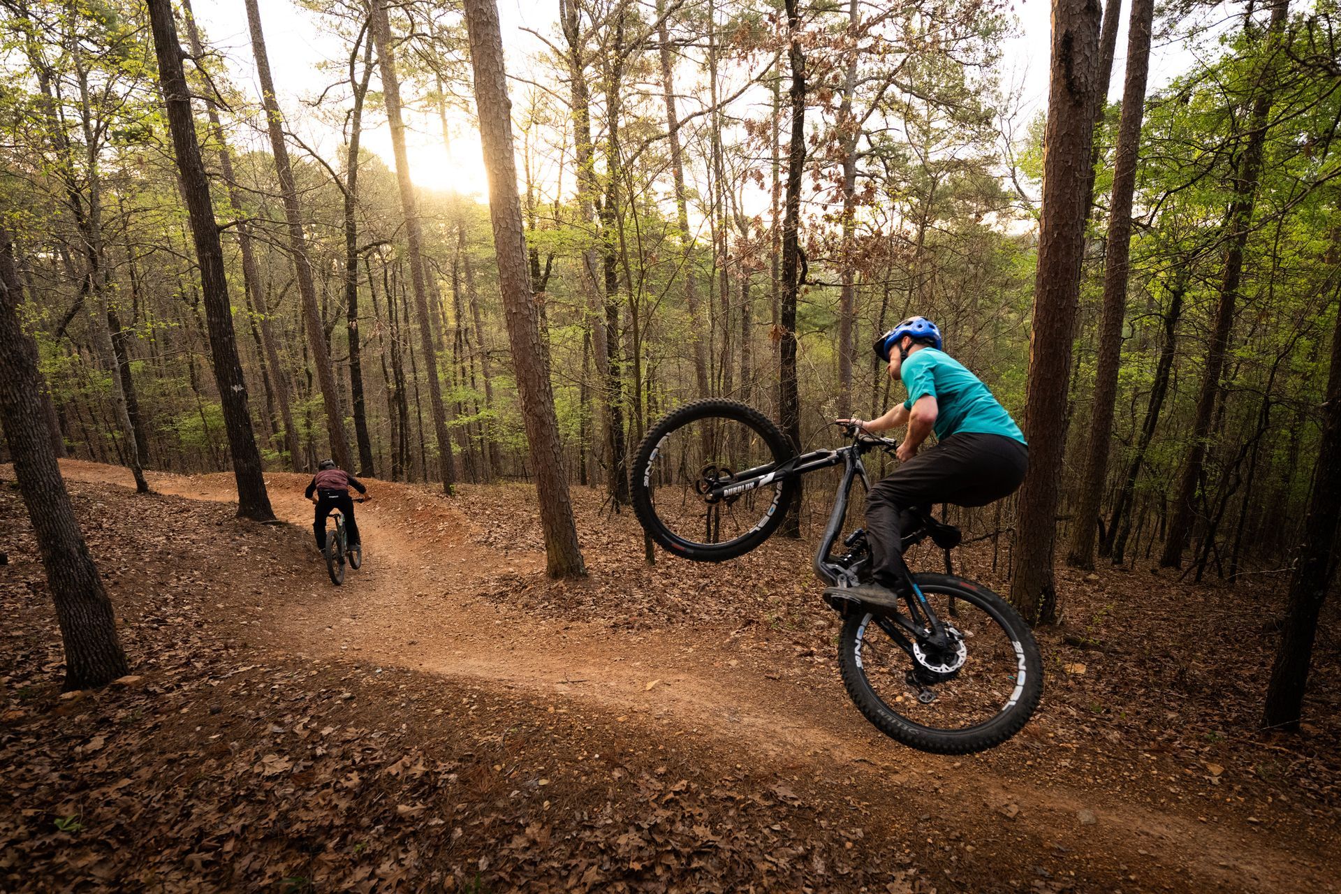 Two mountain bikers on a dirt trail in a forest; one doing a wheelie, the other following.