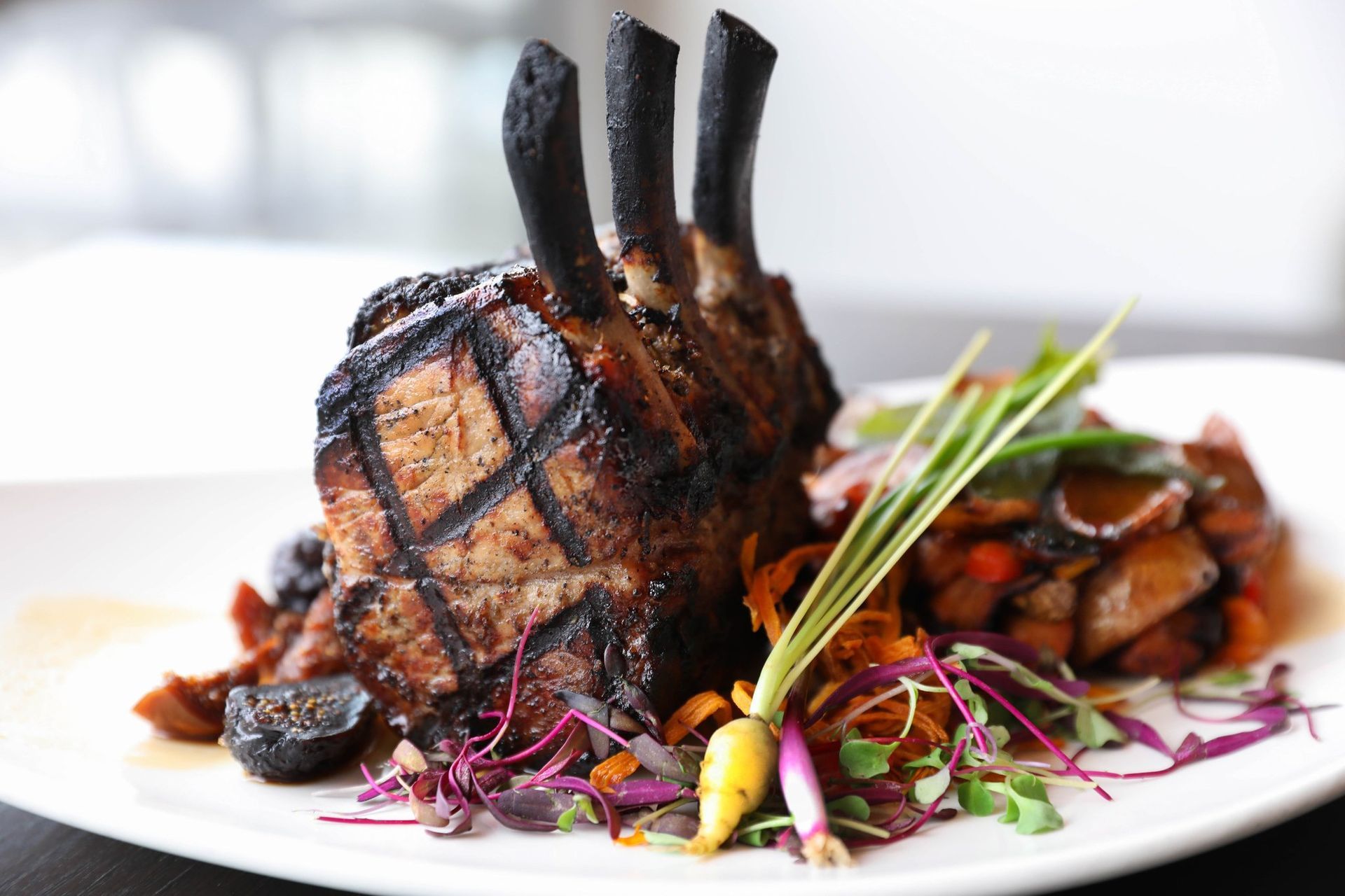 Grilled rack of pork with charred bones, served with vegetables and greens on a white plate.