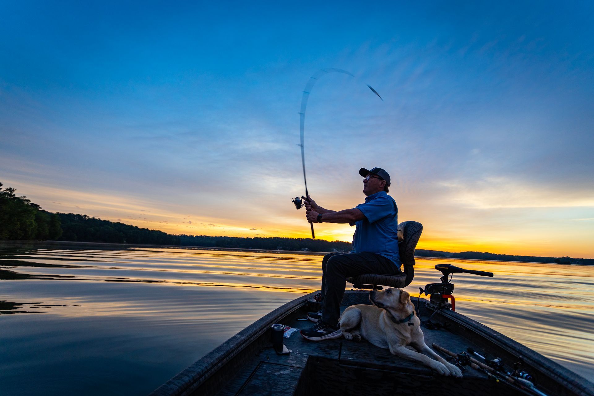 Man fishing on a boat at sunset, dog lying in the front, with blue and orange hues reflecting on the water.
