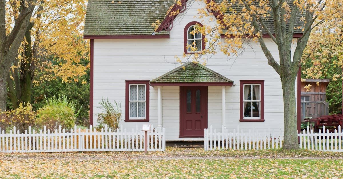 White house with burgundy trim, picket fence, and fall foliage.