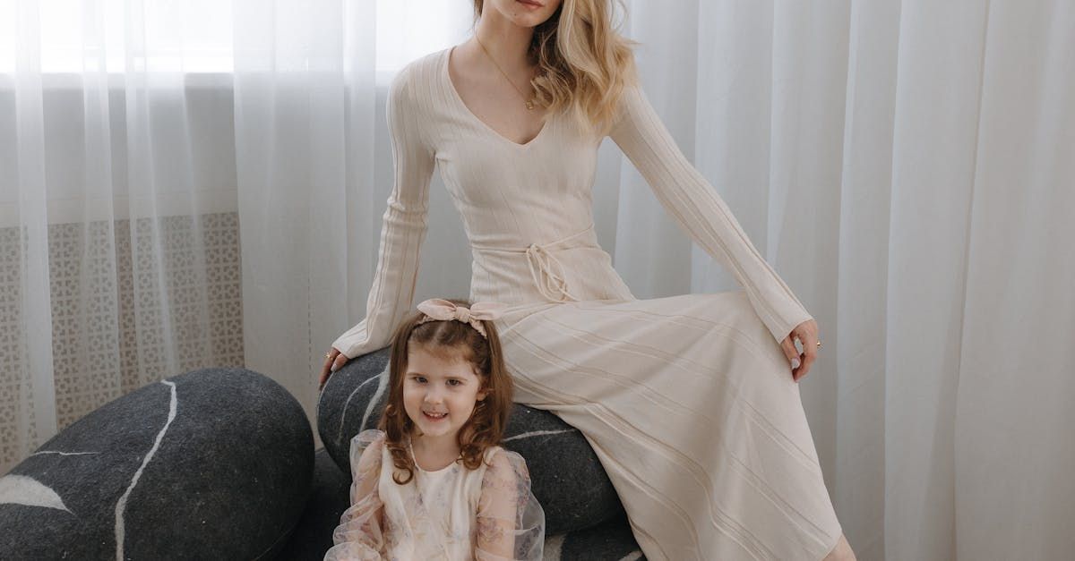 Woman in cream dress sits with young girl, both smiling, in front of a window with white curtains.
