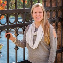 Blonde woman in gray sweater smiles near a black metal gate, fall leaves in the background.