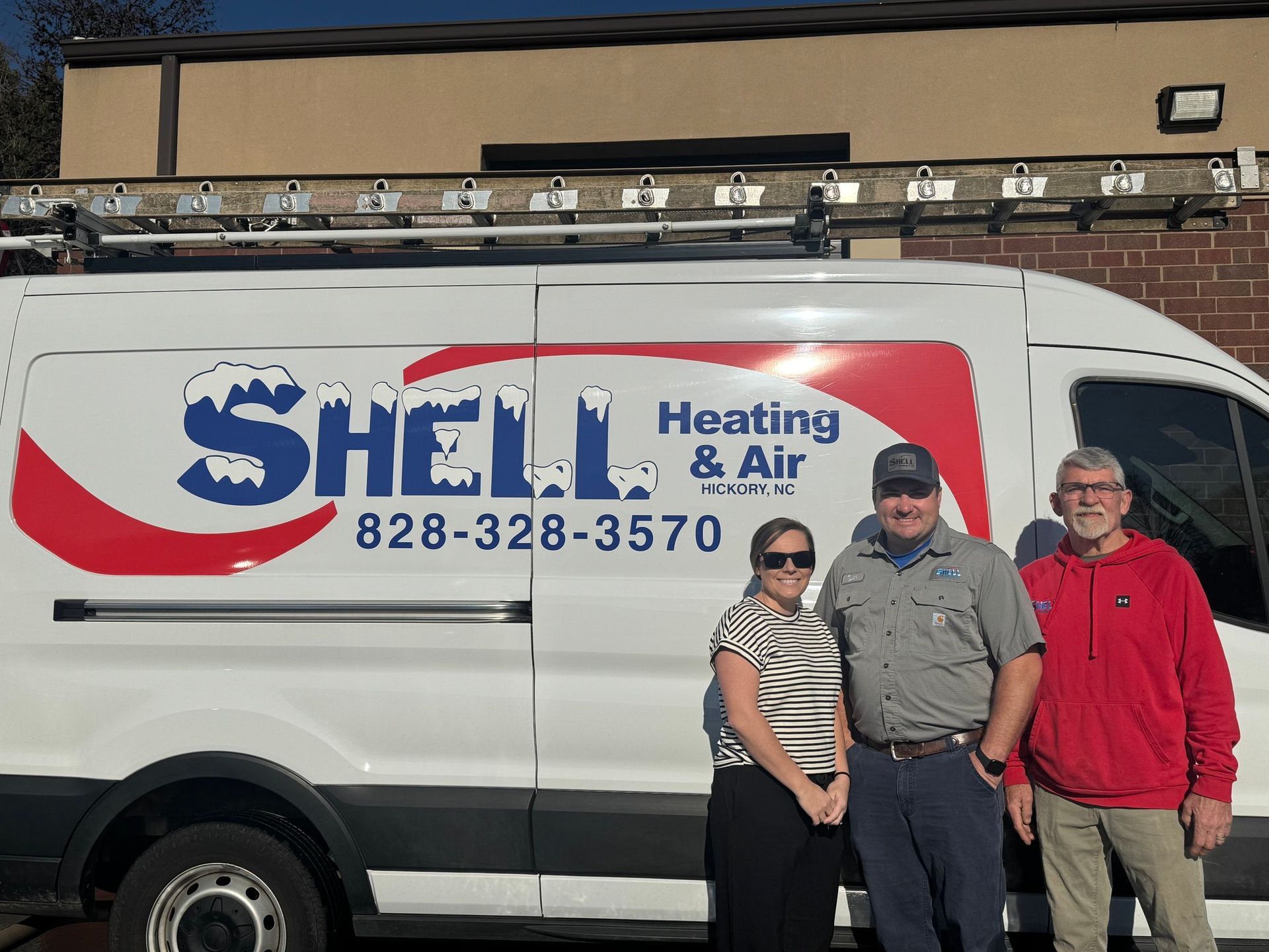 Three people stand smiling in front of a white Shell Heating & Air van with a ladder on top.