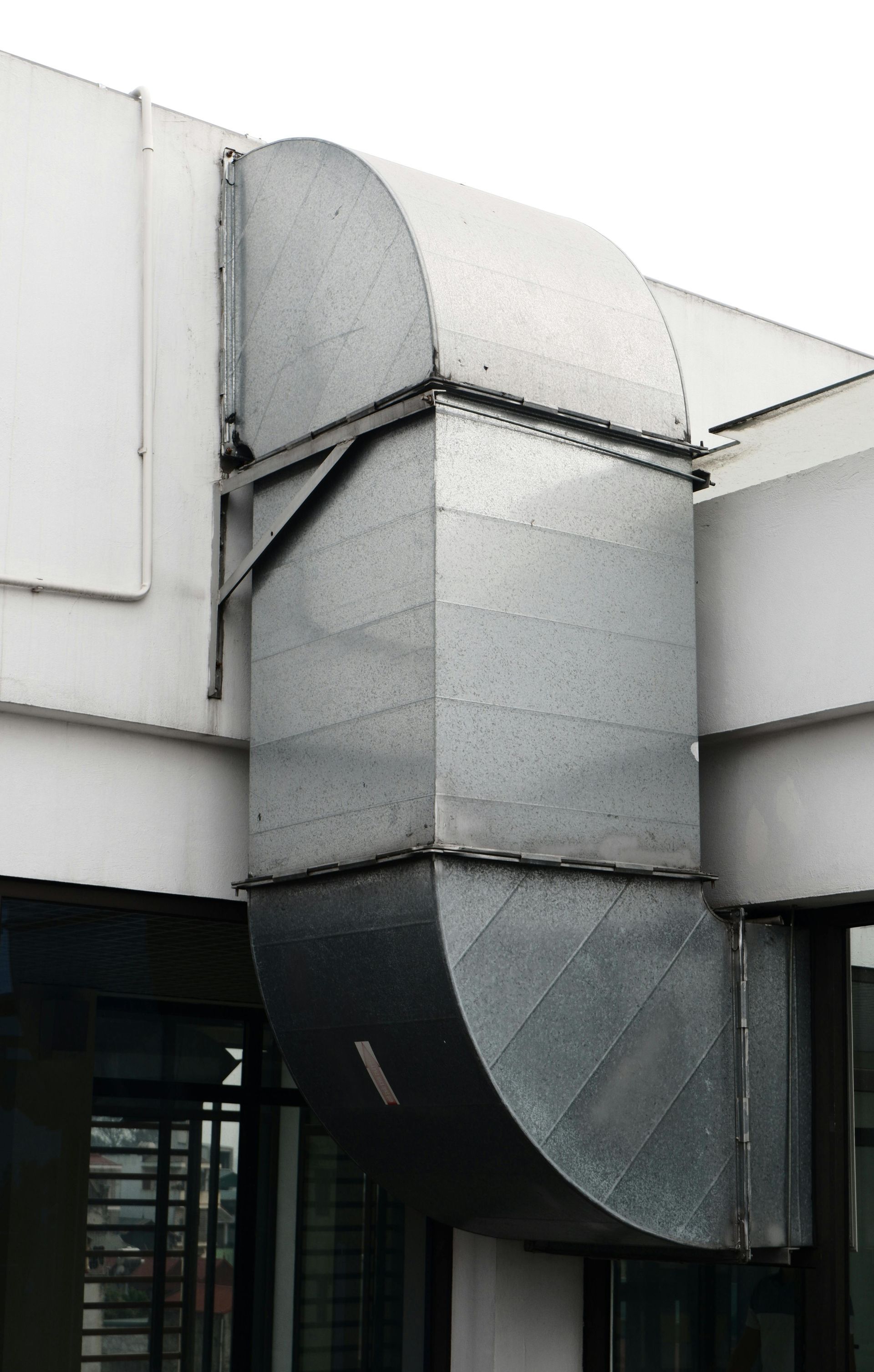 A vertical, galvanized steel HVAC ventilation duct mounted on the exterior white wall of a building.