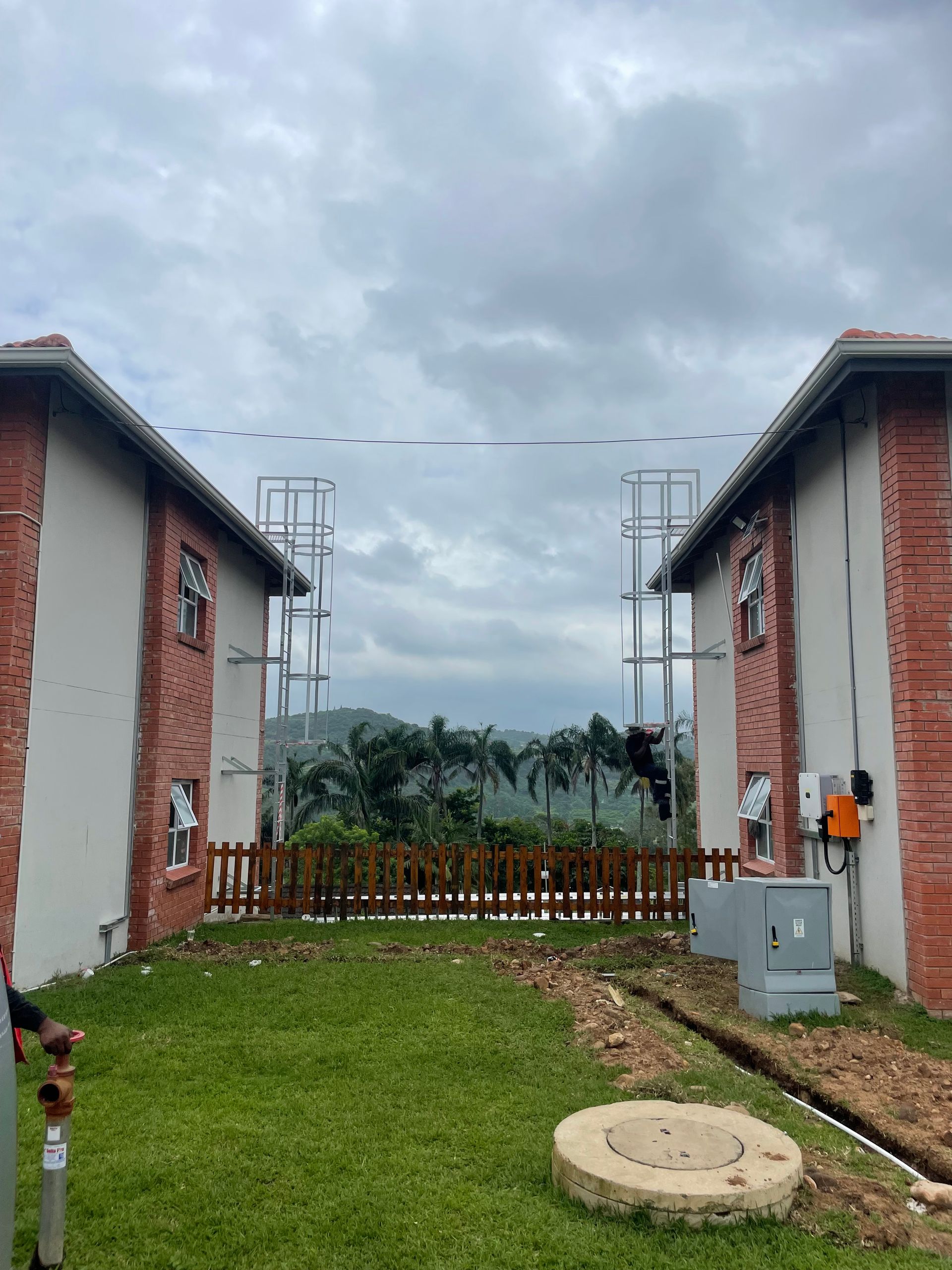 Two brick buildings flank a grassy yard. A wooden fence and trees are visible in the distance under a cloudy sky.