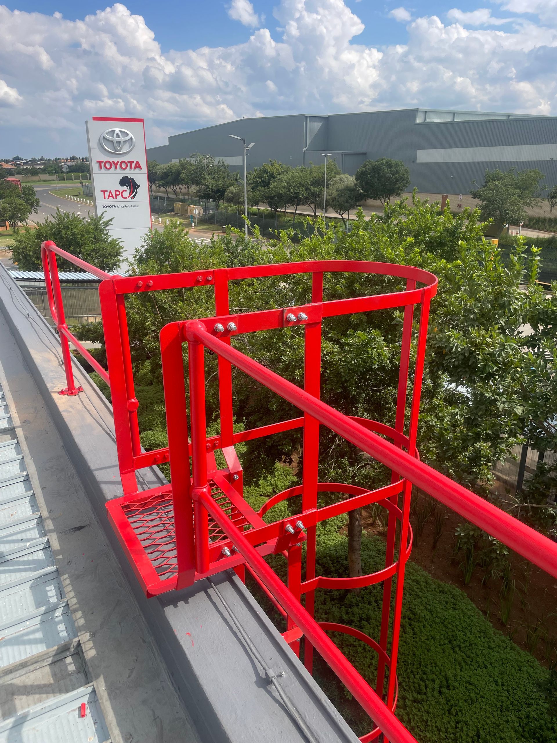 Red safety railing on a rooftop overlooking a Toyota dealership and lush greenery under a cloudy sky.