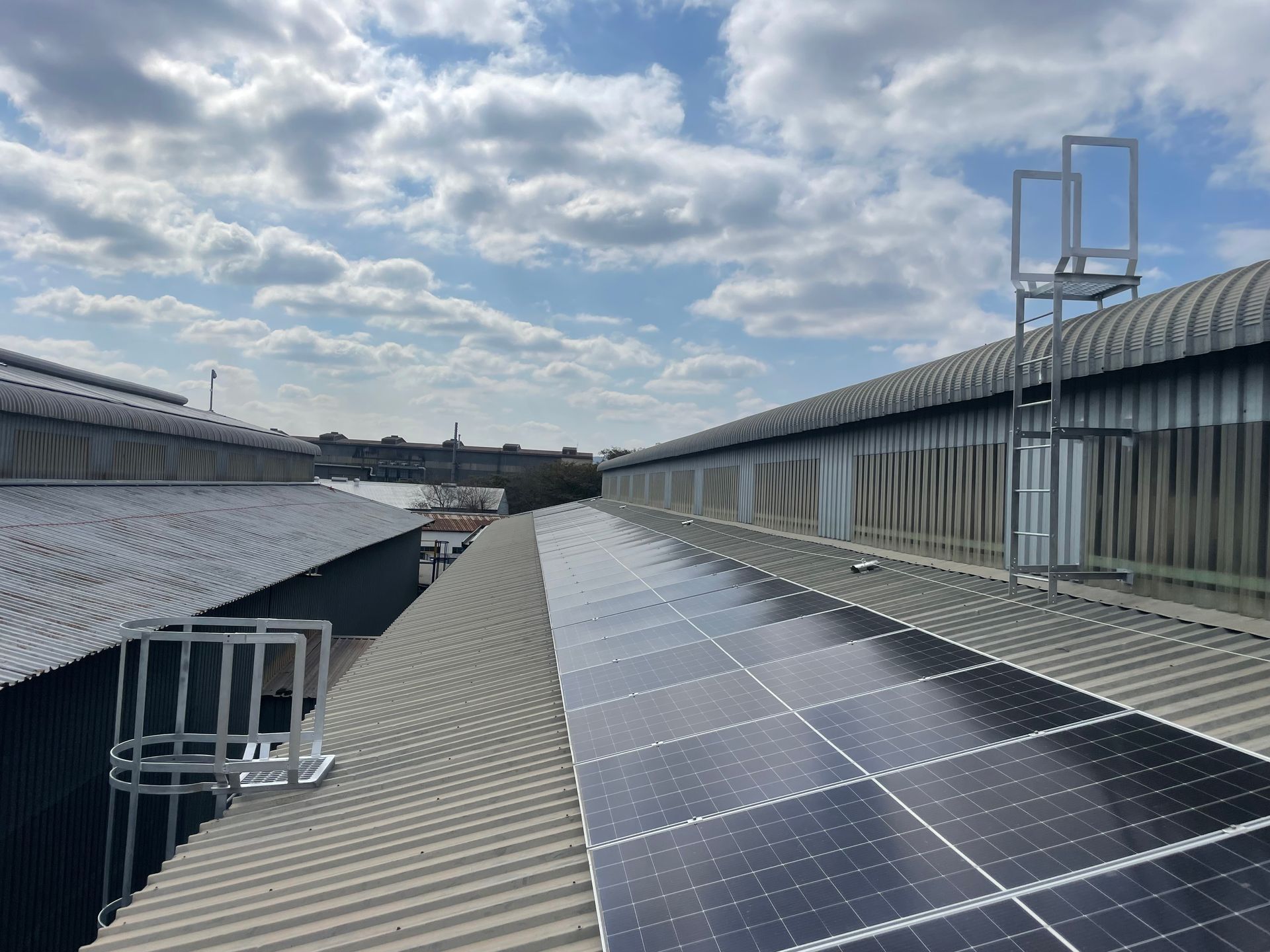Rooftop with solar panels, safety cages, and a ladder, under a cloudy sky.