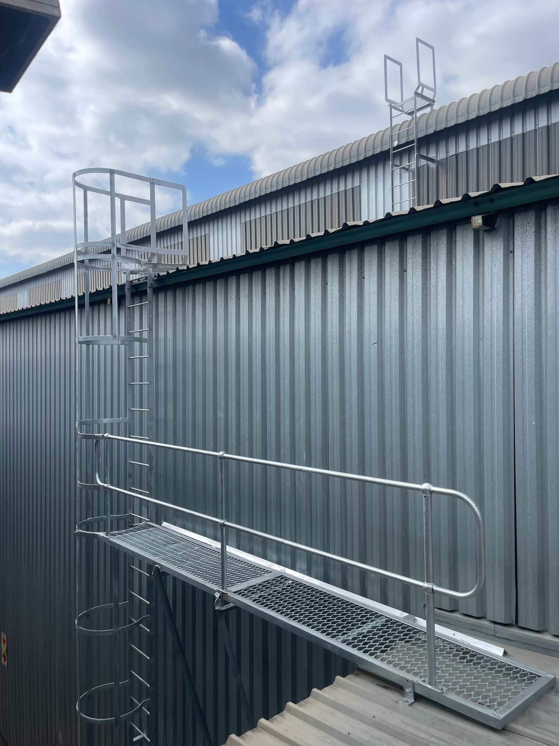Metal ladder with safety cage, platform, and handrails on a corrugated metal building roof.