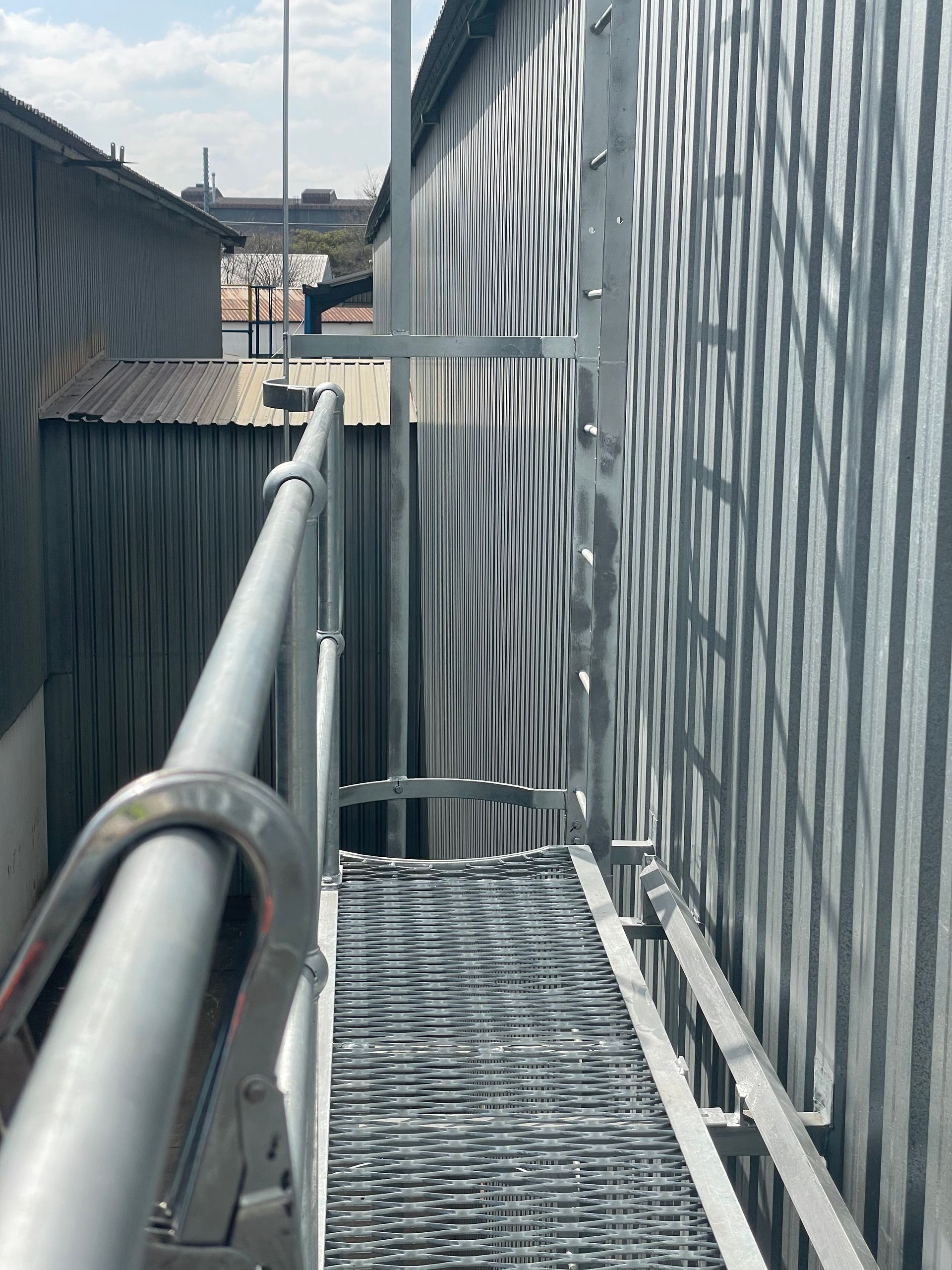 Metal catwalk with handrails and grating leads alongside a corrugated metal building under a blue sky.