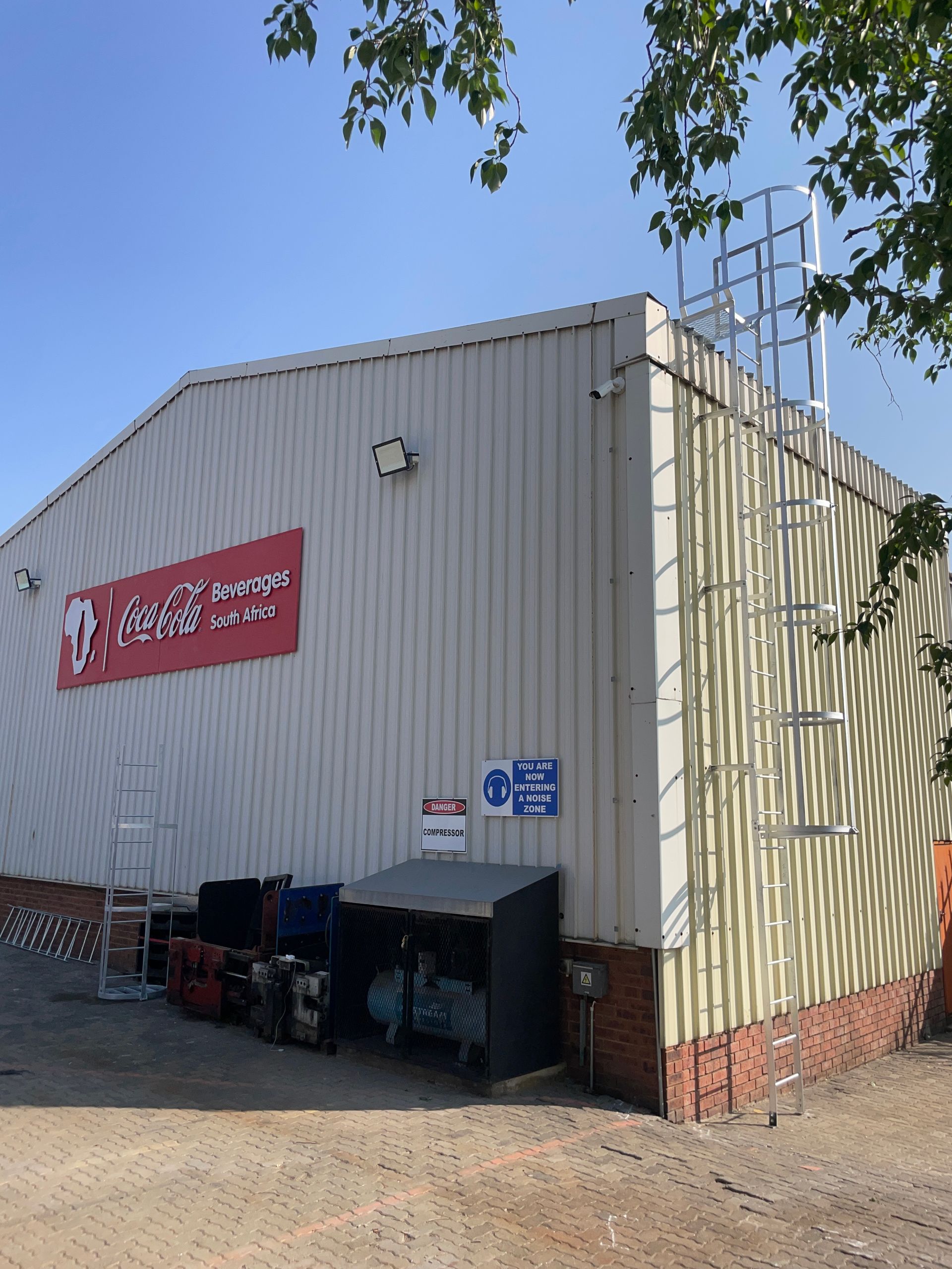 Warehouse with red sign, ladders, and metal siding. Clear sky, brick base, and equipment at the ground level.