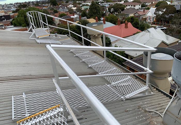 Metal walkway with railing on a rooftop, providing access near chimneys; houses in the background.